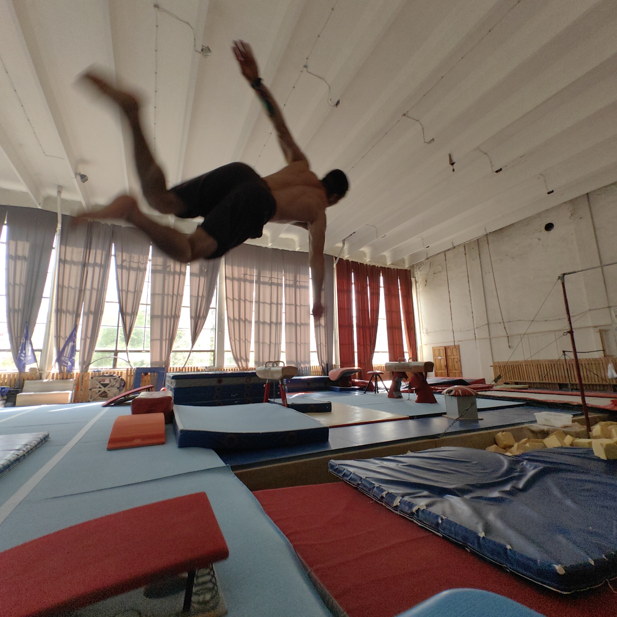 A gymnast performs a mid-air trick inside a gymnasium. The gymnasium appears to be located in Varna, Bulgaria, based on the banners hanging near the windows. The gymnast is male, shirtless, wearing dark shorts, with a tattoo visible on his upper left arm. He is suspended in the air, limbs outstretched, against the backdrop of a large, well-lit gymnasium. Several gymnastics apparatus such as pommel horses, mats, and blocks are present on the floor. Large windows with light-colored curtains line the walls. The ceiling is white with visible wiring and lighting fixtures. The overall image is slightly blurred, indicating movement.