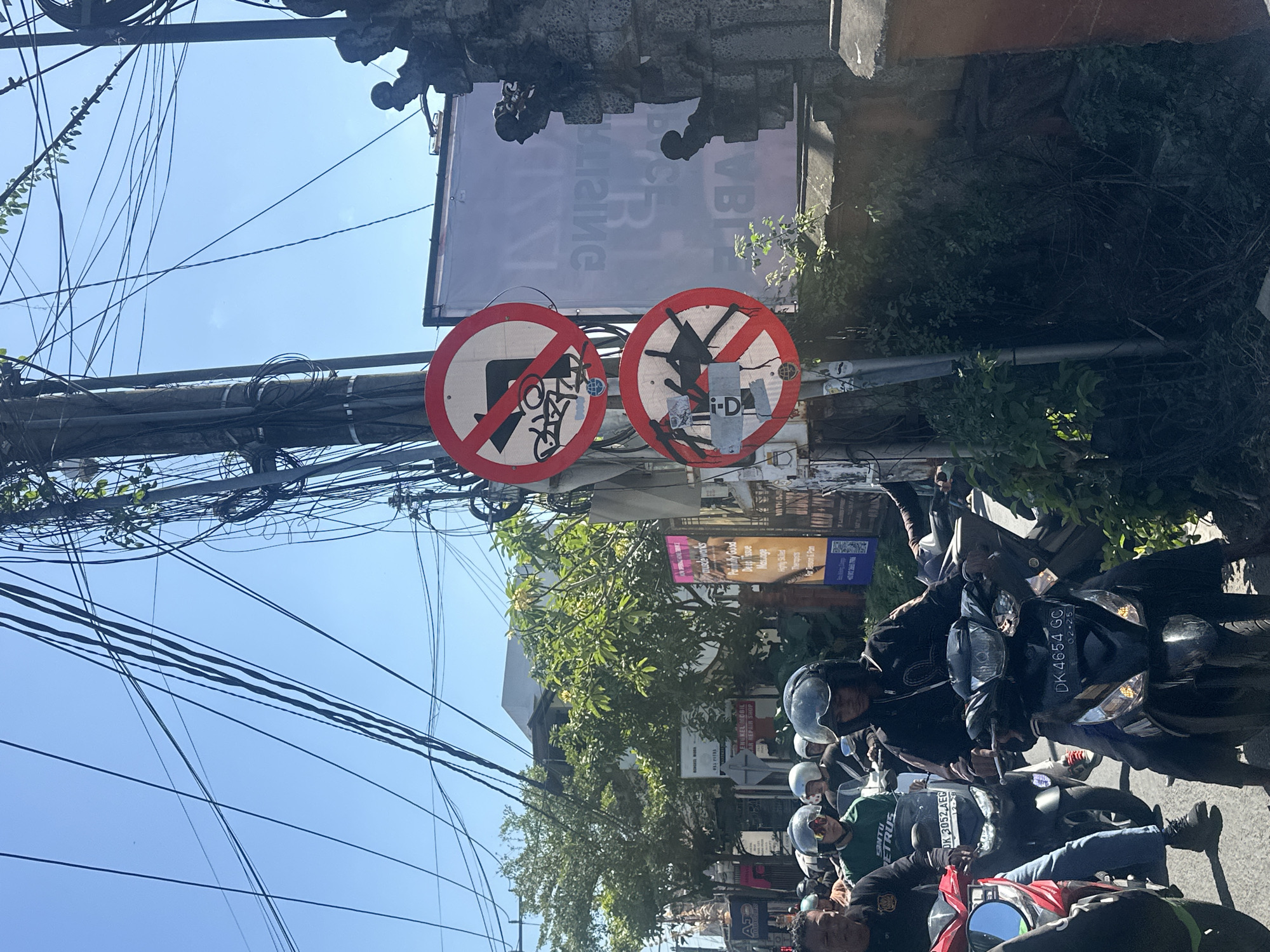 In North Kuta, Indonesia, a street scene unfolds under a clear blue sky, heavily populated with overhead power lines tangled on poles. Traffic, primarily motorcycles, is congested. A group of individuals, mostly wearing helmets, navigate the road on their motorbikes. Two prominent circular red and white prohibition signs are visible on a pole. The top sign depicts a right-turn arrow crossed out, indicating "no right turn." Below it, another sign shows a silhouette of a person on a motorcycle, also crossed out, signifying "no motorcycling." In the background, buildings and lush green trees line the street, interspersed with advertisements and signage. A stone wall with decorative elements stands to the right, contributing to the urban landscape. The atmosphere suggests a busy, everyday moment in a bustling Indonesian city.