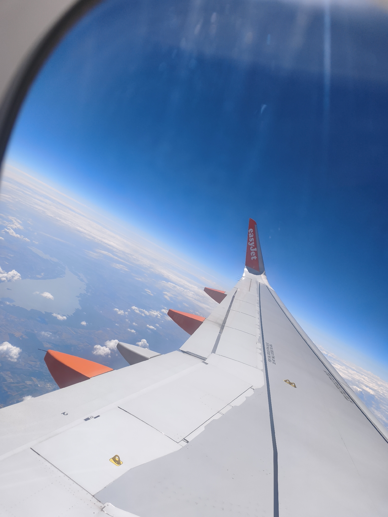 An aerial view from an airplane window shows a vast expanse of blue sky above a layer of white clouds. Below the clouds, the landscape is visible, featuring a large body of water that appears to be a lake or river, bordered by land with agricultural fields. An airplane wing, predominantly white with orange wingtips, extends into the foreground, obscuring part of the view. The wing has the "easyJet" logo printed in red on its upper surface. The image is sharp and clear, capturing the details of the wing and the distant landscape. The angle of the shot suggests the airplane is in flight, likely at cruising altitude. The provided crops offer closer views of the sky, the wing, and the landscape, aiding in identifying specific elements. However, no specific landmarks or identifiable structures within the city of Trebbin, Germany, are discernible from this aerial perspective.