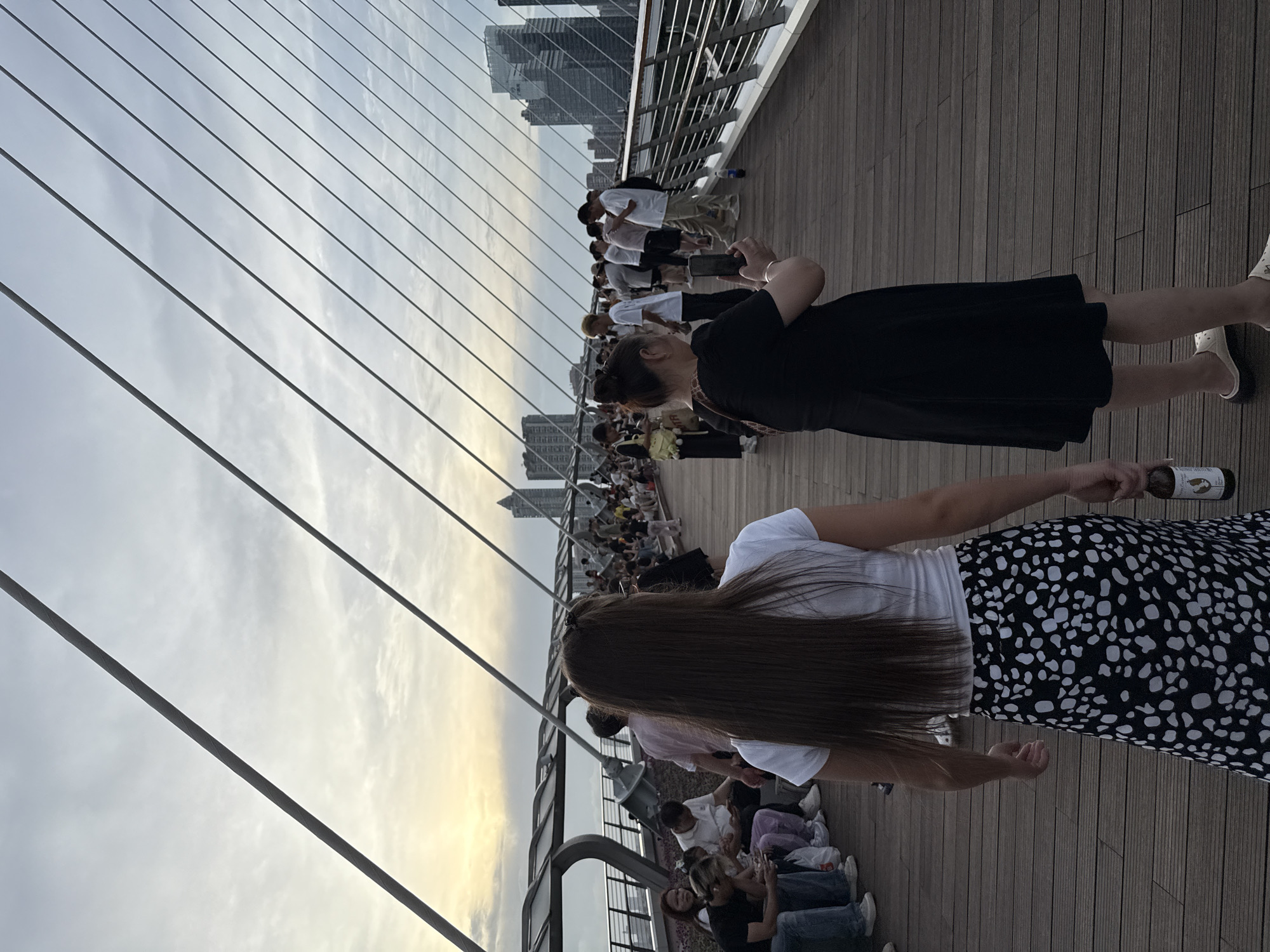 This image depicts a wide shot of a pedestrian bridge at dusk, with numerous people visible walking and sitting. The bridge has a wooden deck and is supported by a network of cables that extend upwards towards a cloudy sky. In the background, modern city buildings are visible, suggesting an urban environment. Several individuals are captured in various activities, including taking photos with their phones and holding drinks. The lighting suggests it is either sunrise or sunset. The scene appears to be within Yuexiu District, China.