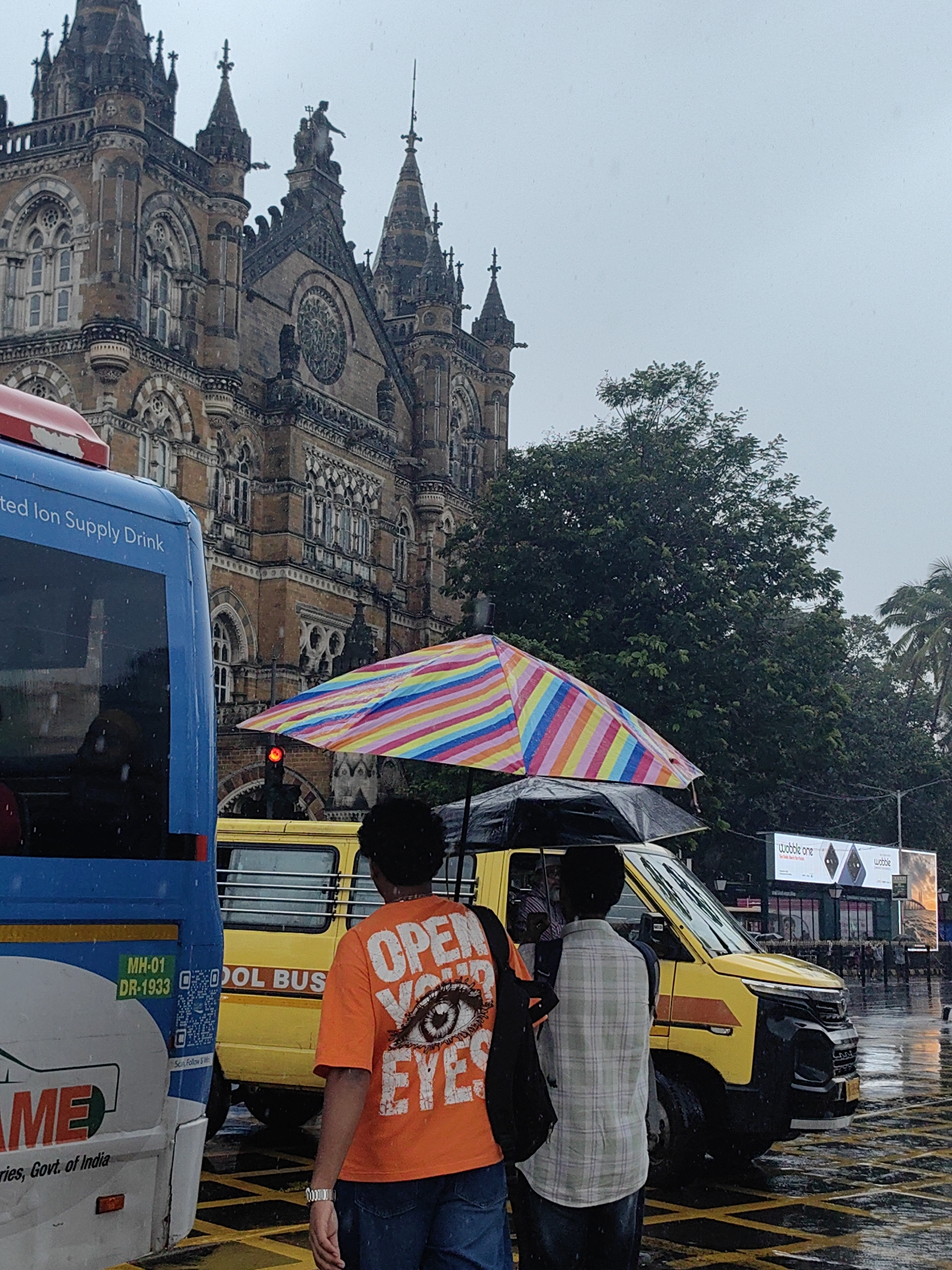 In Mumbai, India, during a rainy day, a street scene unfolds with the grand Victoria Terminus (now Chhatrapati Shivaji Maharaj Terminus) building as a backdrop. The gothic architecture of the historic train station is partially obscured by the overcast sky and the falling rain, which is visible as white streaks in the image. In the foreground, the side of a blue bus is visible, with some text on its side reading "ted Ion Supply Drink." Beside it, a yellow van is parked, with two individuals standing near it. One person, wearing an orange t-shirt with a large eye graphic and the words "OPEN YOUR EYES," is partially visible. Another individual is standing next to them, holding a black umbrella over themselves and the person in the orange shirt, who also has a colorful, striped umbrella. The ground is wet, reflecting the surrounding lights and vehicles, indicating recent or ongoing rainfall. The scene captures a typical urban moment in Mumbai amidst inclement weather.