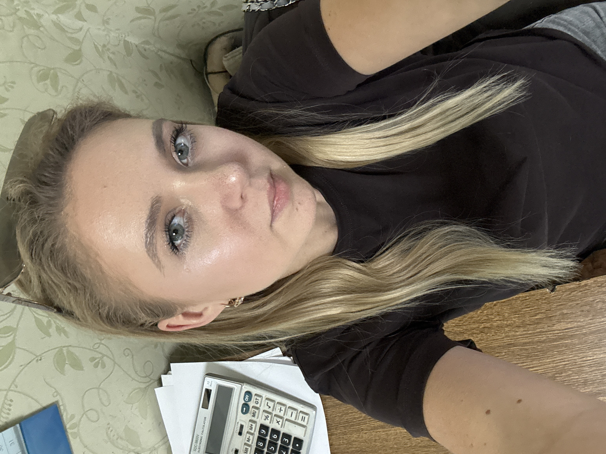 The image shows a close-up, overhead shot of a young woman with blonde hair and blue eyes. She is wearing a dark brown t-shirt and has light makeup on, including mascara and lipstick. Her hair is long and falls across her face and shoulder. She is lying down, with her head slightly tilted.
In the foreground, partially visible, is a white calculator with a black keypad. Some papers are also present beneath it. The background consists of a light green surface with a subtle floral pattern. The overall lighting suggests it could be daytime, but the exact time of day or weather conditions are not discernible. The woman's expression is neutral. There is some visible text on the calculator, including "SDC-9800V ELECTRONIC CALCULATOR" and numerical digits and symbols.