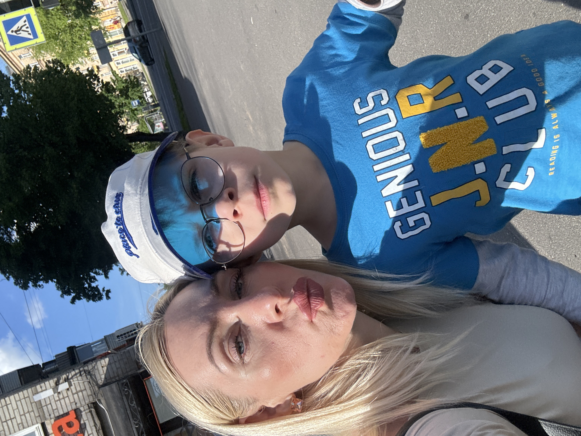 This outdoor selfie features an adult woman and a child, likely a boy, captured in bright daylight under a clear blue sky. The image is significantly tilted, giving a dynamic angle.
The woman, with long blonde hair, is positioned closer to the camera, wearing a light top and sporting full lips in a slight pout. A star-shaped earring is visible on her left ear. Resting against her is the child, who has short hair and round, blue-tinted glasses. He wears a vibrant blue long-sleeved shirt with lighter grey sleeves and a white visor with blue details, which partially shows the text "Place to be" in script.
The child's blue shirt displays prominent text: "GENIOUS J.N.R. CLUB" in a combination of white and textured yellow letters. Below this, smaller white text reads, "READING IS ALWAYS A GOOD IDEA!". Both subjects are looking towards the camera, with the child maintaining a neutral expression.
The background reveals an urban or suburban setting with a paved street or sidewalk, dense green trees, and distant buildings. A distinctive blue and yellow pedestrian crossing sign, common in Russia, is clearly visible in the upper left corner, serving as a location cue. The strong sunlight creates well-defined shadows, indicating a sunny day, likely during the late morning or afternoon. On a building in the lower left, a red "P" or part of a Cyrillic letter is visible.