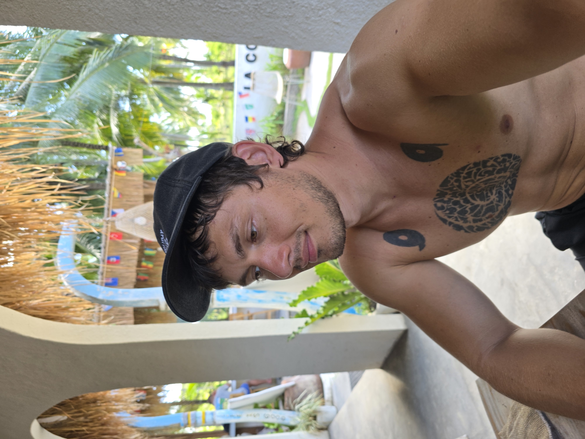 The image is a medium shot of a shirtless man with tattoos, wearing a black baseball cap. He is looking down with a slight smile. The location appears to be outdoors in Nusa Penida, Indonesia, based on the tropical vegetation and the "HOLIDAY" sign visible in the background. The background includes surfboards, a building structure with bamboo-like elements, and lush greenery including palm trees. The light suggests it is daytime.