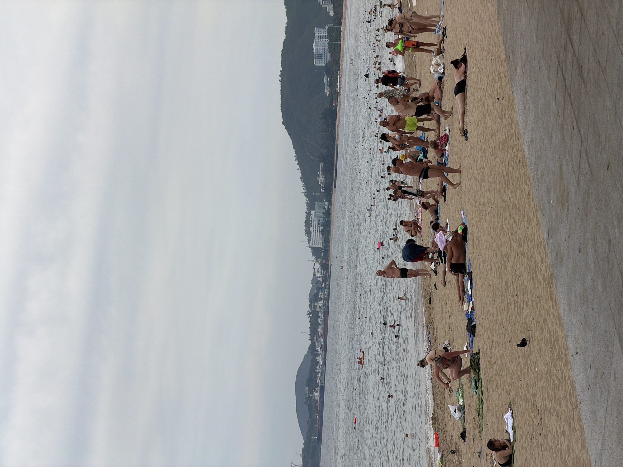 The image depicts a crowded beach in Nha Trang, Vietnam, under an overcast sky. Numerous people are engaged in various beach activities: swimming in the ocean, sunbathing on the sand, and socializing. The ocean is populated with swimmers, and a small boat with two individuals is visible. In the background, hills and buildings line the coastline, suggesting an urban setting. The beach itself is sandy, with towels and beach bags scattered across it. A few birds are present on the sand. The overall scene conveys a busy day at a popular beach destination.