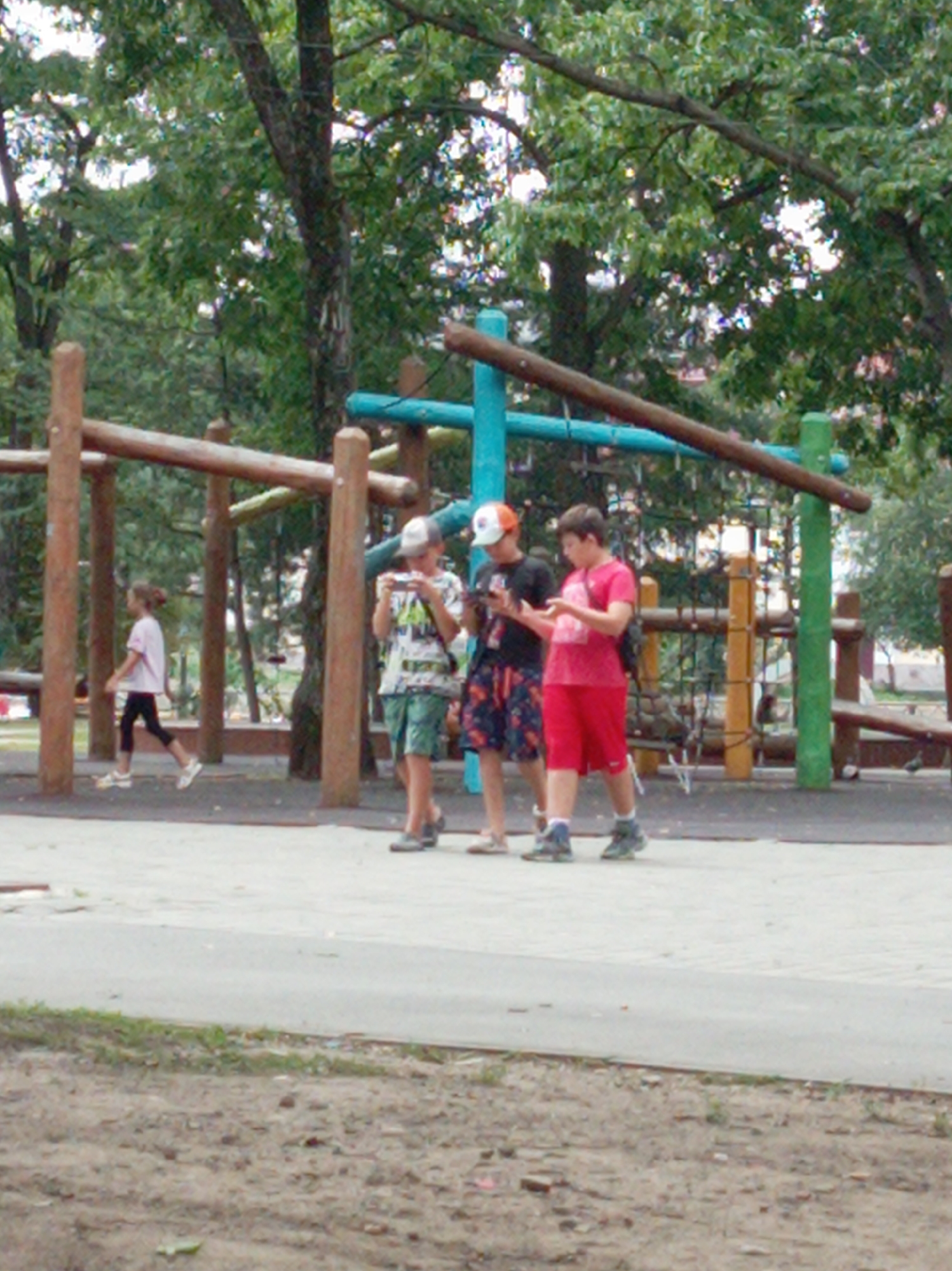 The image captures a scene at a playground in Artyom, Russia. In the foreground, a paved surface transitions to a dirt area. A wooden playground structure with horizontal bars and vertical posts is visible in the mid-ground. A young girl with brown hair tied in a bun, wearing a pink t-shirt and black leggings, walks towards the left of the frame. To her right, three boys are present. One boy, wearing a white and black patterned t-shirt, green shorts, and a baseball cap, is looking at his phone. Beside him, a boy in a red t-shirt and red shorts is also holding a phone. Further to the right, another boy in a black t-shirt and patterned shorts is partially visible, also looking at a phone. The background consists of trees and foliage, indicating an outdoor setting. The overall lighting suggests it is daytime, likely overcast.
