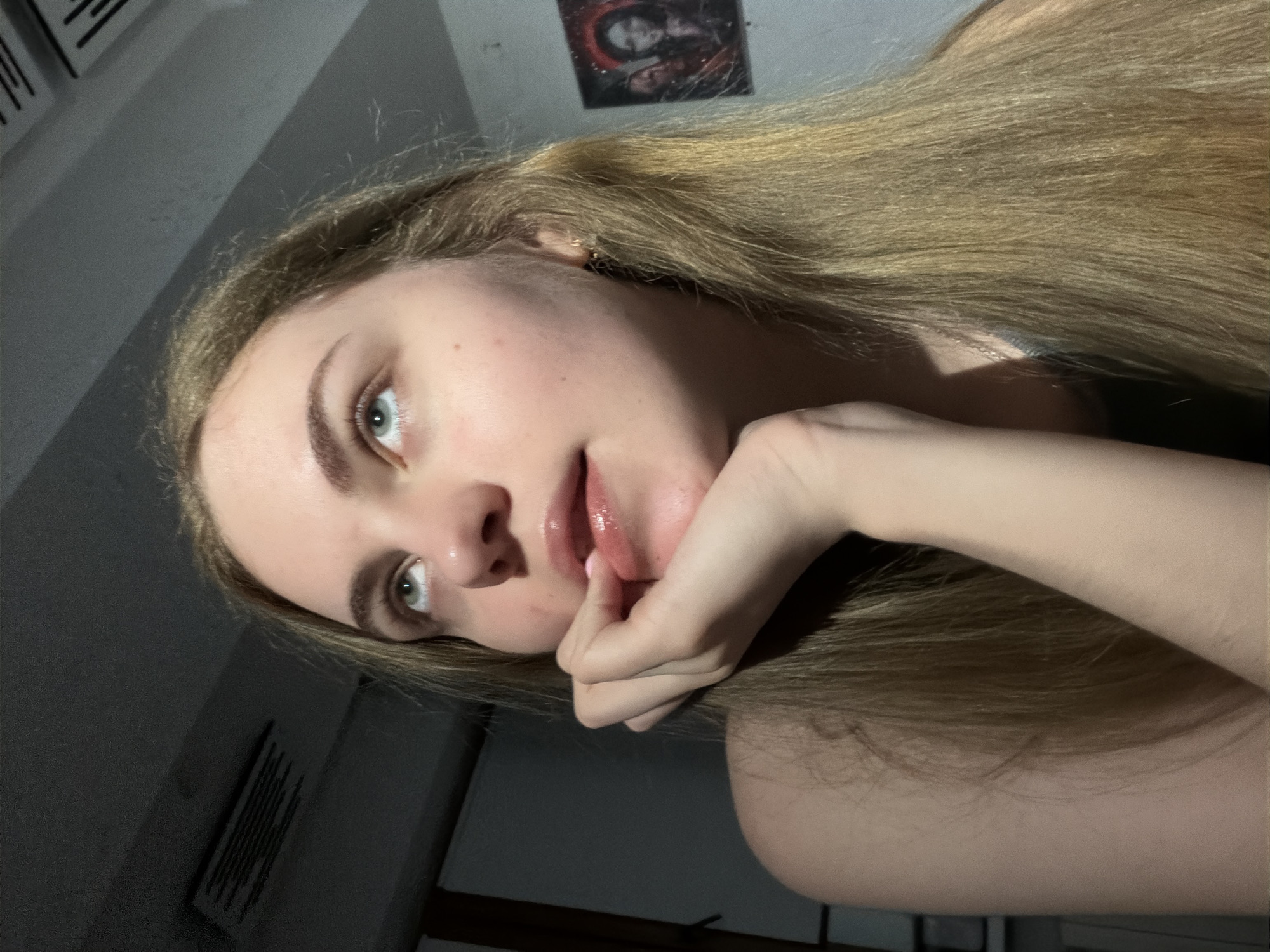 The image shows a close-up of a young woman's face, with her head tilted upwards and her gaze directed towards the upper left corner of the frame. Her right hand is placed under her chin, with her thumb and index finger gently touching her lips. She has long, blonde hair that falls around her shoulders. The lighting in the image is dim, with a strong light source casting shadows on her face and the surrounding environment. In the background, a framed picture can be seen on the wall, which appears to depict two figures. The overall setting suggests an indoor environment, possibly a residential space. There is no indication of the specific location within the city of Dnipro, Ukraine.