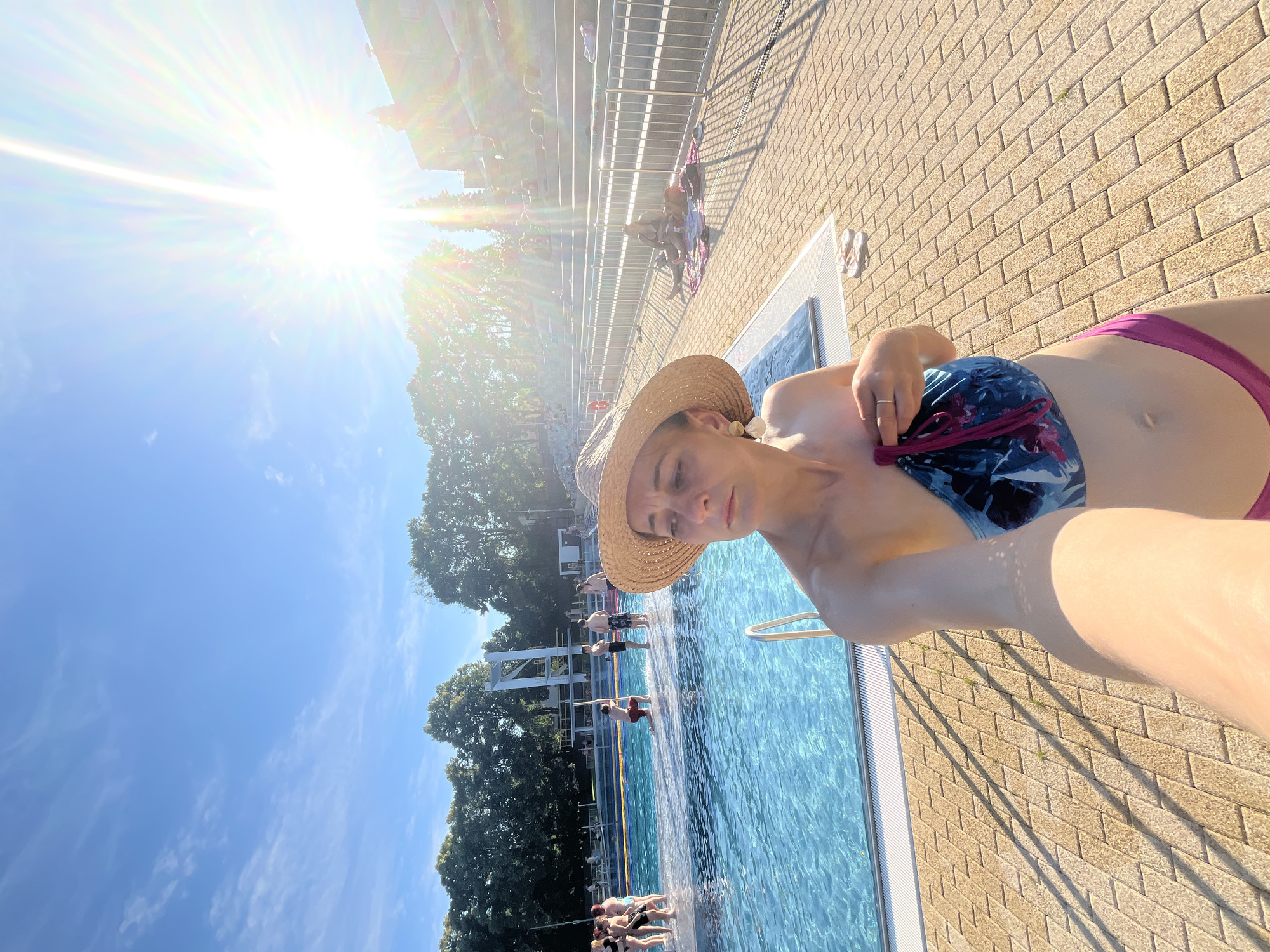 A woman wearing a straw hat and a bikini stands in the foreground of the image, holding a colorful swimsuit. She is at a public swimming pool on a sunny day. The pool is filled with clear blue water, and several people are visible in and around it. Some are swimming, while others are relaxing on lounge chairs. The background shows a building with tiered seating and trees. The sun is shining brightly, creating lens flare and casting shadows on the tiled ground.