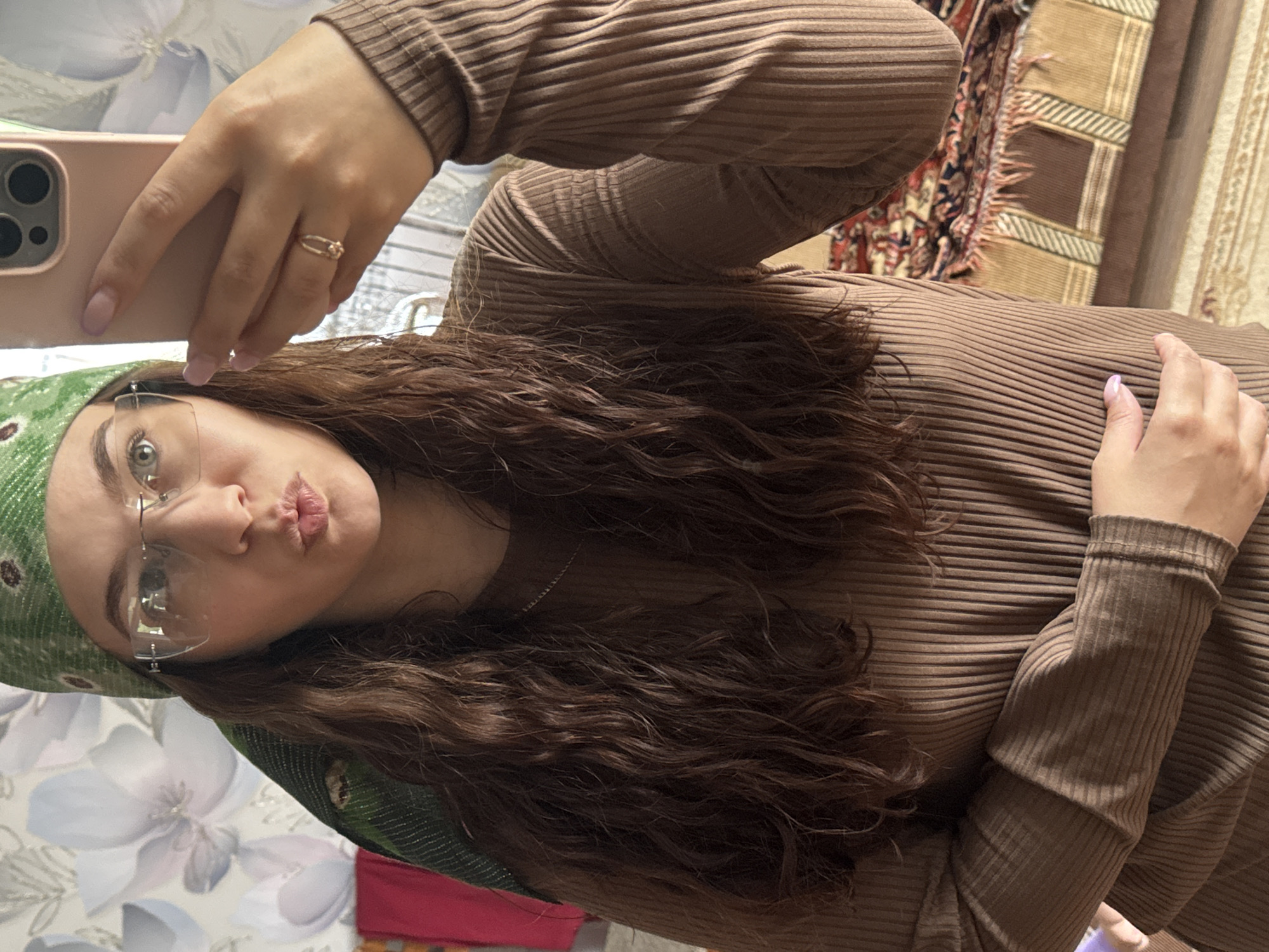A woman is taking a selfie. She is wearing a brown ribbed top and a green headscarf. She has long brown curly hair, glasses, and is wearing a ring. The background appears to be a floral-patterned surface and part of a patterned rug or throw. There is no evidence to confirm the location in the city of Nizhnyaya Gar', Russia.