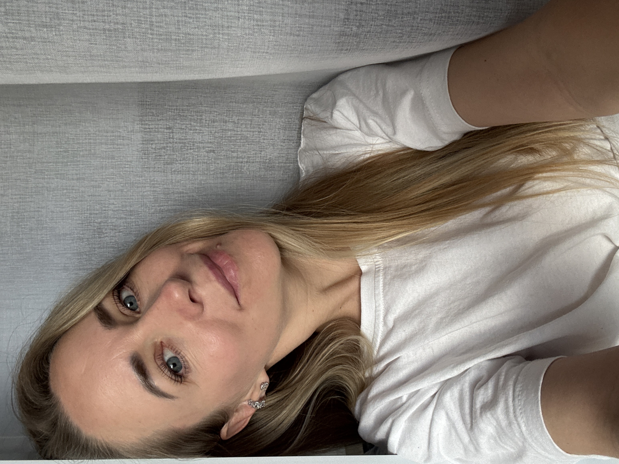 The image shows a close-up, waist-up selfie of a Caucasian woman with long blonde hair and blue eyes. She is wearing a white t-shirt and silver stud earrings. She is leaning against a textured gray background, possibly a wall or curtain. The lighting is soft and diffused. The scene appears to be indoors. There is no indication of the city of Chaikovsky, Russia, or any specific urban environment.