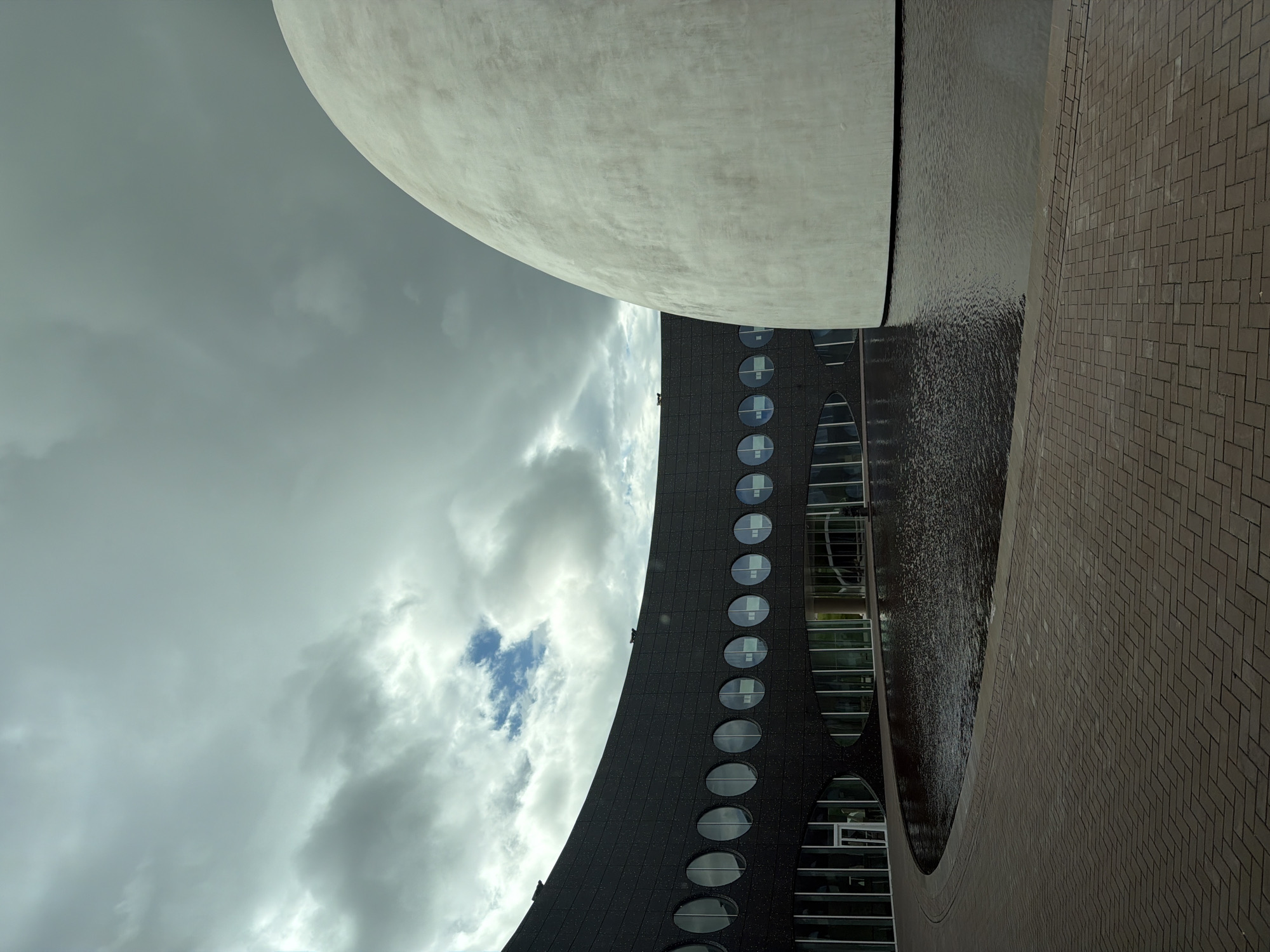 The image depicts the exterior of the Yelizovo Planetarium, a distinctive spherical building located in Yelizovo, Russia. The planetarium is characterized by its large, white dome and a curved, dark-colored building with a series of circular windows. In front of the buildings, there is a body of water reflecting the overcast sky. The foreground consists of a paved walkway made of brick pavers arranged in a herringbone pattern. The sky is filled with heavy, gray clouds, suggesting cloudy weather. No people are visible in the scene.