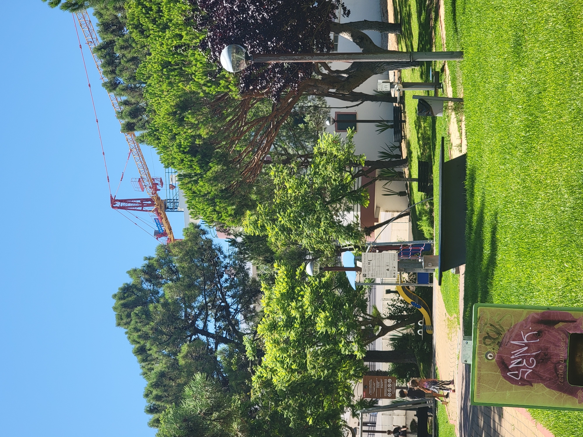 This is an eye-level, medium-shot photograph of a park in Quarteira, Portugal, on a clear, sunny day. In the background, a construction crane with red and yellow elements is visible against a bright blue sky, suggesting ongoing development in the area. The park features lush green grass, mature trees with dense foliage, and paved pathways.
In the middle ground, there is a playground with a yellow slide and a climbing structure. Two children, a boy and a girl, are walking on the pathway near the playground. A dark-colored bench is positioned on the grass in the foreground. Several lampposts are present throughout the park, including one with a spherical light fixture.
To the left of the frame, a green rectangular bin with graffiti art is prominent. The graffiti depicts a stylized figure with white lettering, partially obscuring the bin's surface. Signs with text and graphics are also visible near the playground area, likely providing information or rules for park users. The overall scene conveys a sense of a public recreational space during daytime.