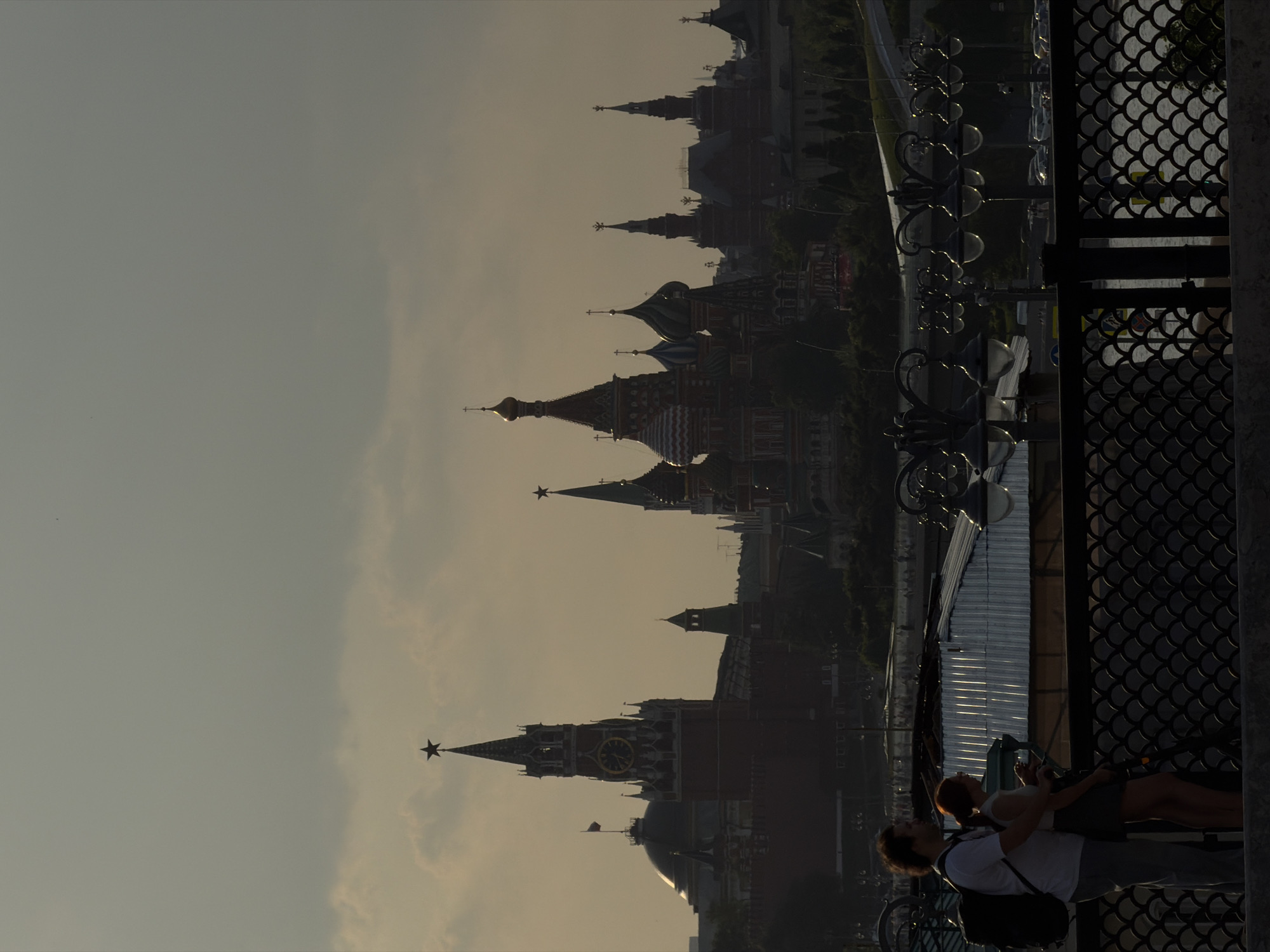 A photo captures the Moscow Kremlin in Russia at dusk. The Kremlin's spires, domes, and clock tower are silhouetted against a gray sky. In the foreground, two people are seen next to a wrought-iron fence, their figures obscured by the low light.