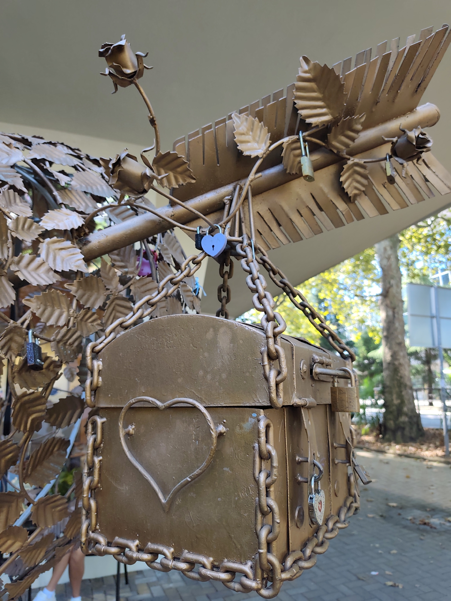 The image captures a close-up of a golden metallic sculpture in the city of Sochi, Russia, designed to resemble a treasure chest adorned with roses, leaves, and an arrow. The chest is heavily chained and secured with multiple padlocks, including a small blue heart-shaped padlock and a larger brass padlock. The sculpture is part of a decorative outdoor installation, likely a popular spot for couples to attach "love locks." In the background, sunlight filters through trees, suggesting an outdoor setting on a clear day. The ground is paved with interlocking stones, and fallen leaves indicate the season may be autumn. A person's legs, wearing white sneakers, are partially visible on the left side of the frame, suggesting the sculpture is accessible to the public. The overall impression is one of romantic symbolism and public art.