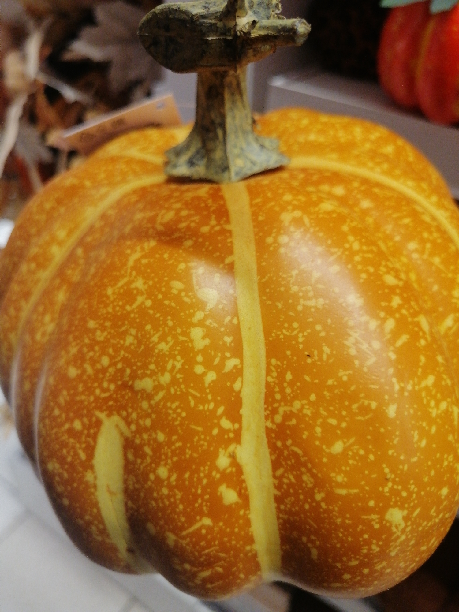 The image shows a close-up of a decorative, orange pumpkin with yellow speckles and vertical yellow stripes. The pumpkin has a brown, textured stem. In the background, out of focus, are dried leaves and another object that appears to be a red, decorative pumpkin or gourd. The scene is indoors, likely a retail environment or home decoration setting. There is no indication of the location being Kremenchuk, Ukraine.