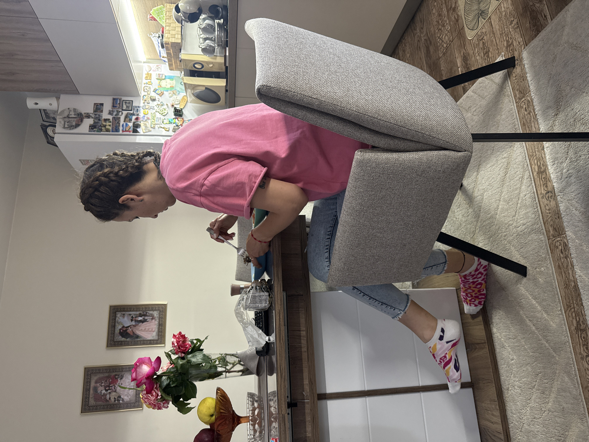 A woman with braided hair, wearing a pink t-shirt and blue jeans, sits on a grey chair with black legs. She is hunched over, working on something on a wooden table. On the table is a vase of red and pink roses, and a fruit bowl with an apple and other fruits. The location appears to be a home in Elhovo, Bulgaria. The background includes pictures on the wall and a refrigerator with magnets and photos. The floor is covered with a patterned rug. The woman is using a fork-like tool. The scene is domestic and suggests an everyday activity.