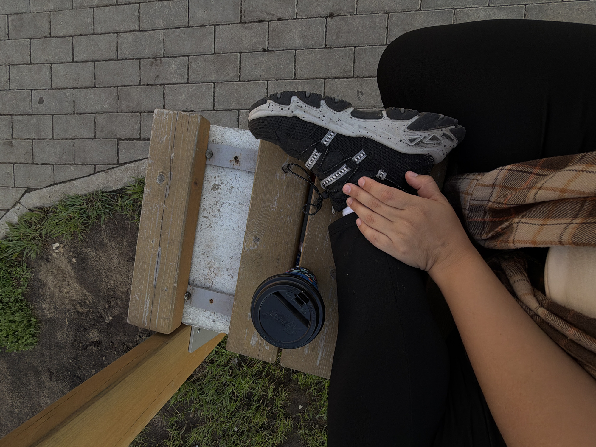 A person is sitting on a wooden bench outdoors, their legs extended and crossed at the ankle. They are wearing black pants and a black athletic shoe with a speckled grey sole. A black disposable coffee cup with a lid sits on the bench to the left of the person's legs. The bench is made of weathered wood and is supported by metal brackets. The ground around the bench consists of dirt and grass on the left, and a grey brick pathway on the right. The scene appears to be in a park or public outdoor area. The image was taken in Froly, Russia.