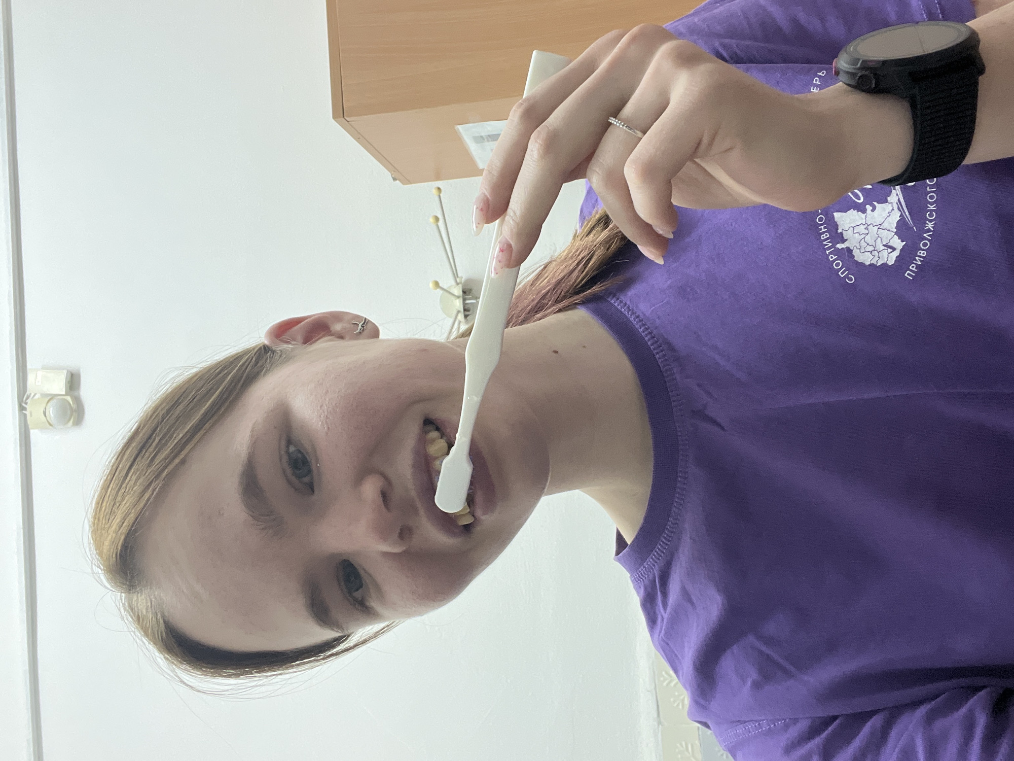 A woman is brushing her teeth with a white toothbrush. She is wearing a purple t-shirt with a graphic and text in Russian. She also has a black smartwatch on her left wrist. The background appears to be indoors, with a white wall and a cabinet. It is not possible to determine if this is in the city of Berezniki, Russia.