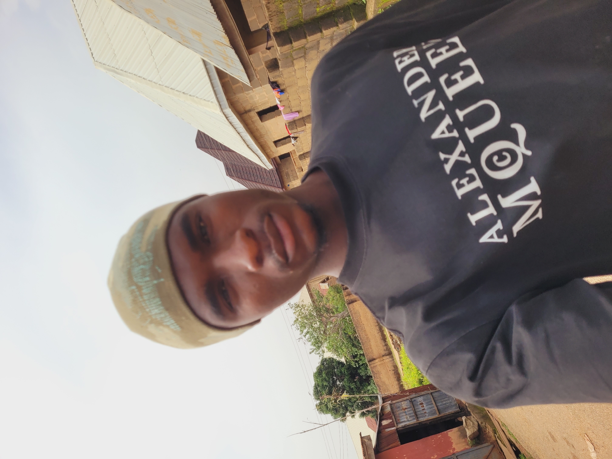 This is a selfie of a young man in Bukuru, Nigeria. The man is wearing a black t-shirt with the words "ALEXANDER MCQUEEN" in white lettering. He is also wearing a light brown beanie. The background shows several houses with corrugated iron roofs and brick walls. There is also some greenery visible. The lighting suggests it is daytime.