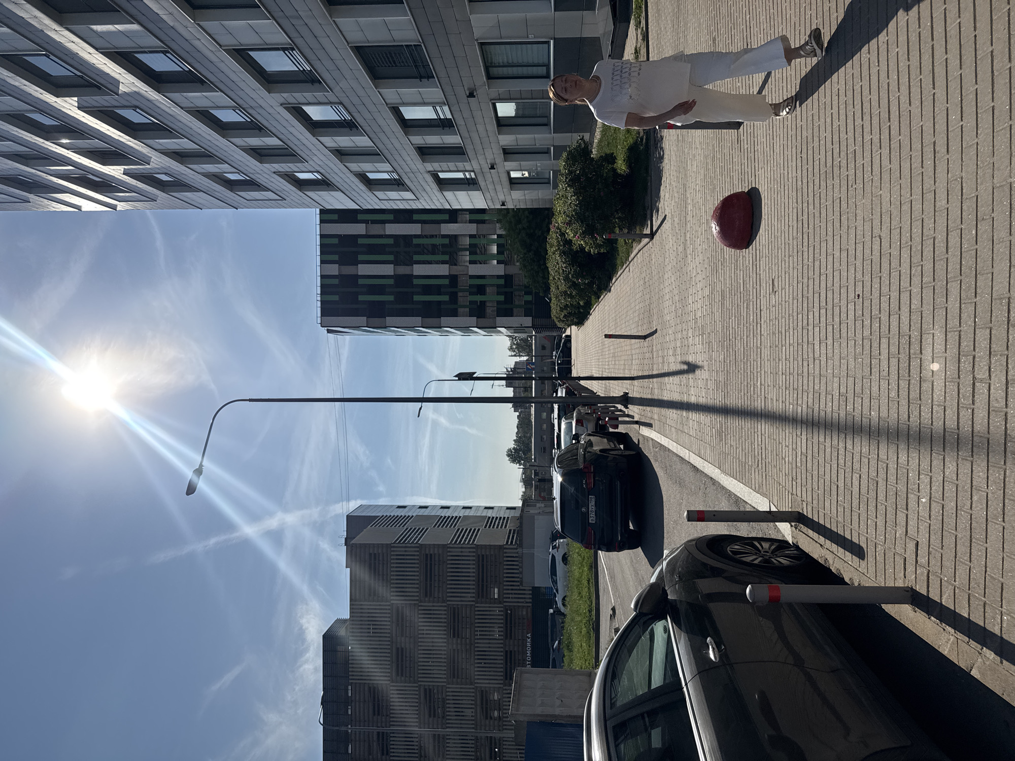 An eye-level, wide shot captures a sunny street scene with modern architecture. In the foreground, a dark-colored car is parked along a sidewalk paved with brick. Two short, grey bollards with red bands stand beside the car. A woman in a white t-shirt and white pants walks away from the camera on the sidewalk. She is wearing sandals and her hair is pulled back. To her left, a large, red, dome-shaped object sits on the sidewalk. Behind the sidewalk, a row of green bushes lines the edge of a grassy area. In the background, two tall, modern buildings stand side-by-side. The building on the left has a facade with a patterned design of horizontal and vertical wooden slats. The building on the right features a dark facade with repeating green and black rectangular panels. Further down the street, a light-colored building with many windows is visible. The sky is a clear blue with thin white contrails from aircraft crisscrossing the sky. A bright sun is visible in the upper left quadrant of the image, casting long shadows from the streetlights and bollards. Several cars are parked or driving on the street.