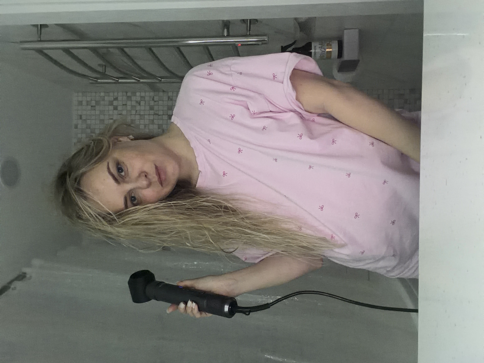 A woman with blonde hair is holding a black electrical massager in her right hand. She is wearing a pink T-shirt with a pattern of red bows and is standing in what appears to be a bathroom or shower stall. Behind her is a towel rack and a tiled wall. The scene is well-lit, and the woman is looking directly at the camera. The specific location in Votkinsk, Russia, cannot be determined from the provided image.