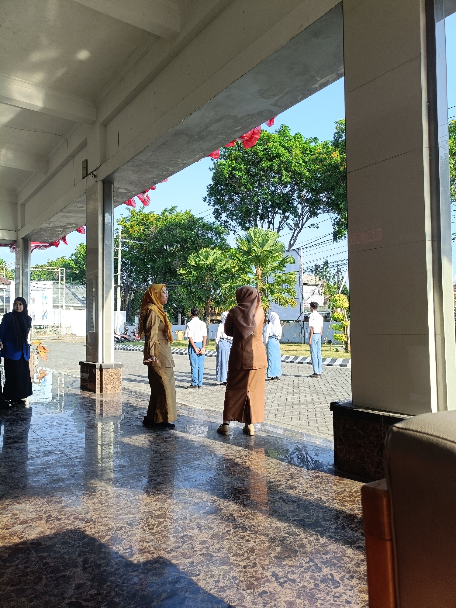 This image depicts a scene outside what appears to be a public building in Kediri City, Indonesia. The foreground is dominated by a highly polished, marbled floor reflecting the light and overhead structure. In the mid-ground, several individuals are visible. Two women in brown uniforms, possibly teachers or staff, are standing under a covered walkway. One woman on the left is looking to her left, while the other on the right is looking forward. Behind them, several students in white shirts and blue pants are lined up outdoors, appearing to be participating in some sort of assembly or event. The background reveals lush green trees, a clear blue sky, and hints of other buildings and electrical wires, indicating an urban or semi-urban environment. Red and white flags are draped along the edge of the overhead structure, suggesting a national celebration or commemoration. The lighting indicates it is daytime, with the sun casting shadows on the reflective floor.