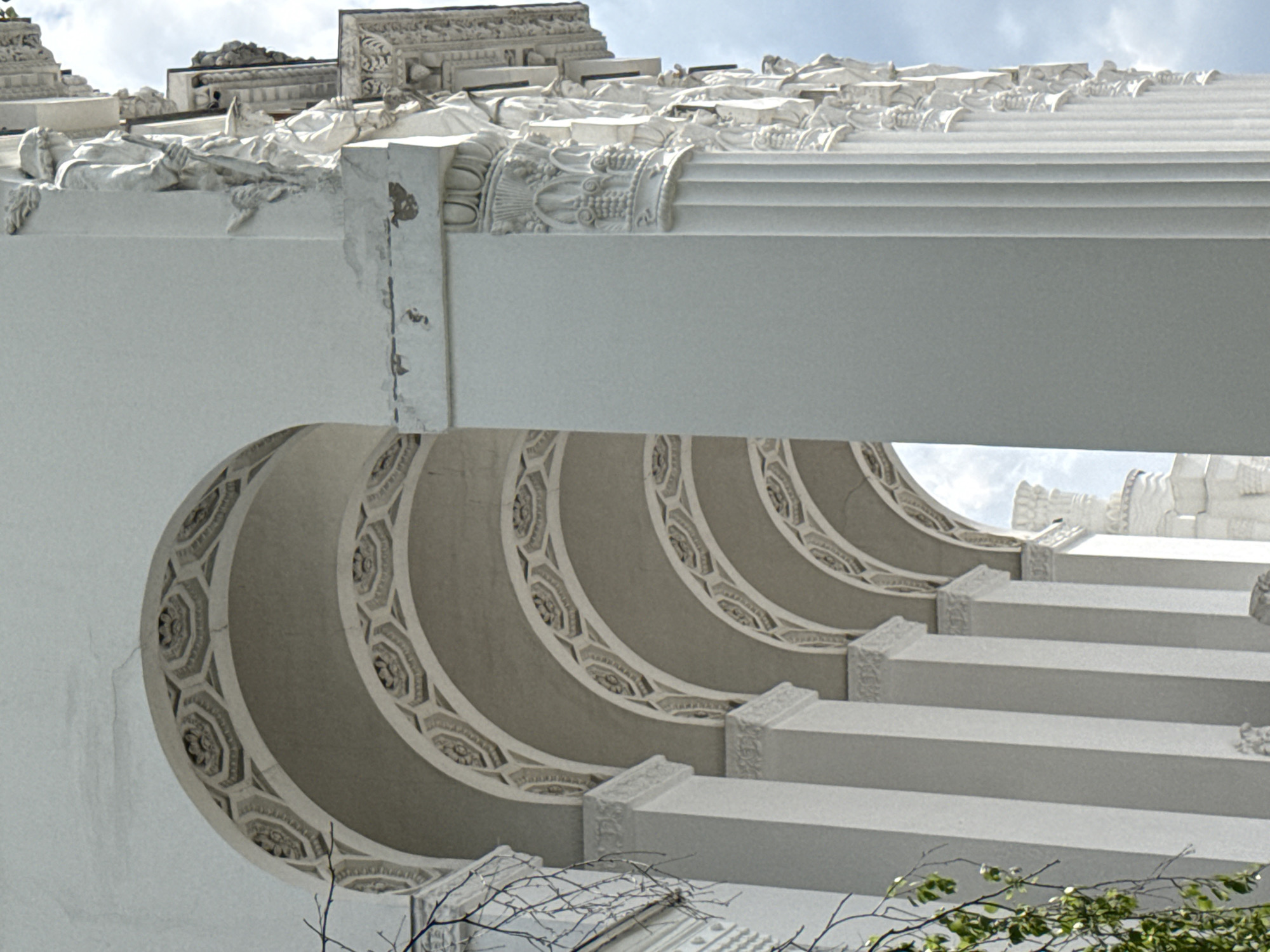 The image depicts a section of the Moscow Kremlin's architectural details, likely a portion of one of its cathedrals or palaces. The scene features white, ornate architectural elements, including a decorated archway with concentric patterned borders, columns, and a detailed roofline with classical motifs and sculptures. The structural components show signs of weathering and age. The sky is visible in the background.
