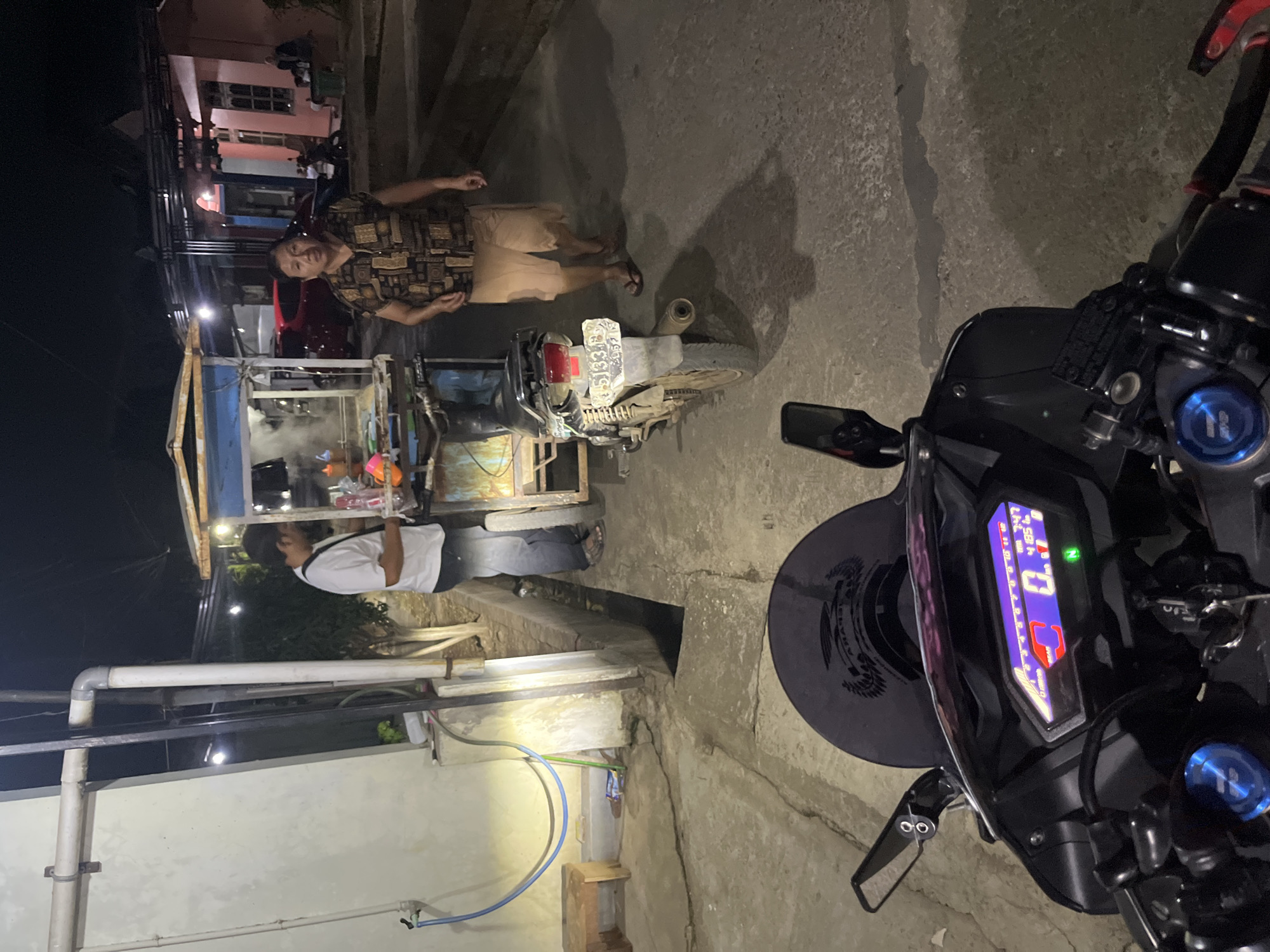 In Karangrandu, Indonesia, at night, a scene unfolds near a food stall. A man in a white t-shirt and jeans stands behind the food cart, which is emitting steam. A woman in a patterned short-sleeved shirt and light brown shorts walks away from the cart towards the right side of the frame. A motorcycle is parked to the left of the woman, facing away from the camera. The foreground is dominated by the handlebars and digital display of a motorcycle, showing the time as 7:47 and a speed of 0. The environment appears to be a paved outdoor area with buildings in the background, illuminated by artificial lighting.