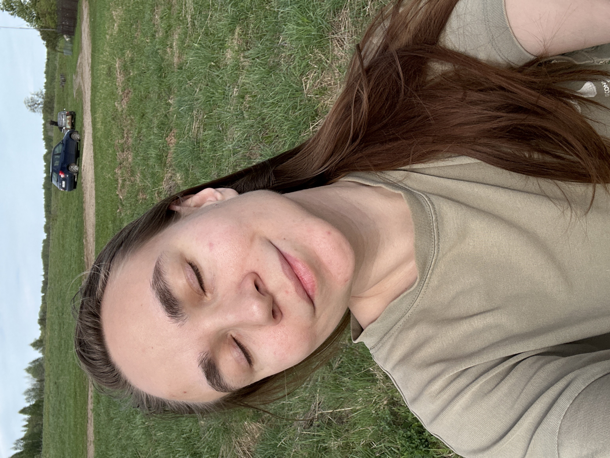 A woman is lying in a field of grass, eyes closed. She has brown hair and is wearing a tan shirt. In the background, there is a dirt path with two vehicles on it. The location is outdoors, likely in a field in the city of Kharnavy, Russia.