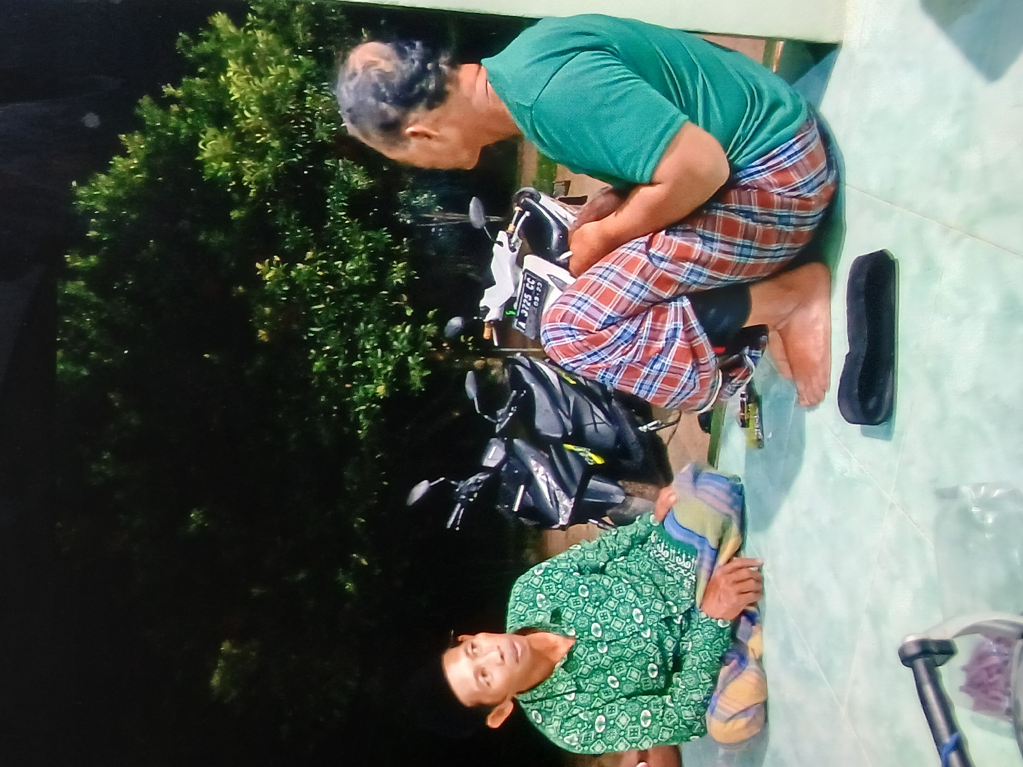 The image shows two men sitting on a tiled floor outdoors at night, with a motorcycle parked behind them. The man on the left is wearing a green shirt with a patterned design and a black cap. He is sitting cross-legged, with a sarong wrapped around his waist. The man on the right is wearing a green t-shirt and plaid pants. He is also sitting, facing away from the camera. The background consists of dark trees and foliage, illuminated by some artificial light, suggesting it is a residential area. A motorcycle is parked between the two men, and a black sandal is visible on the floor in front of the man on the right. The setting appears to be a residential yard in Setia Bakti, Indonesia, likely during an evening gathering or casual conversation.