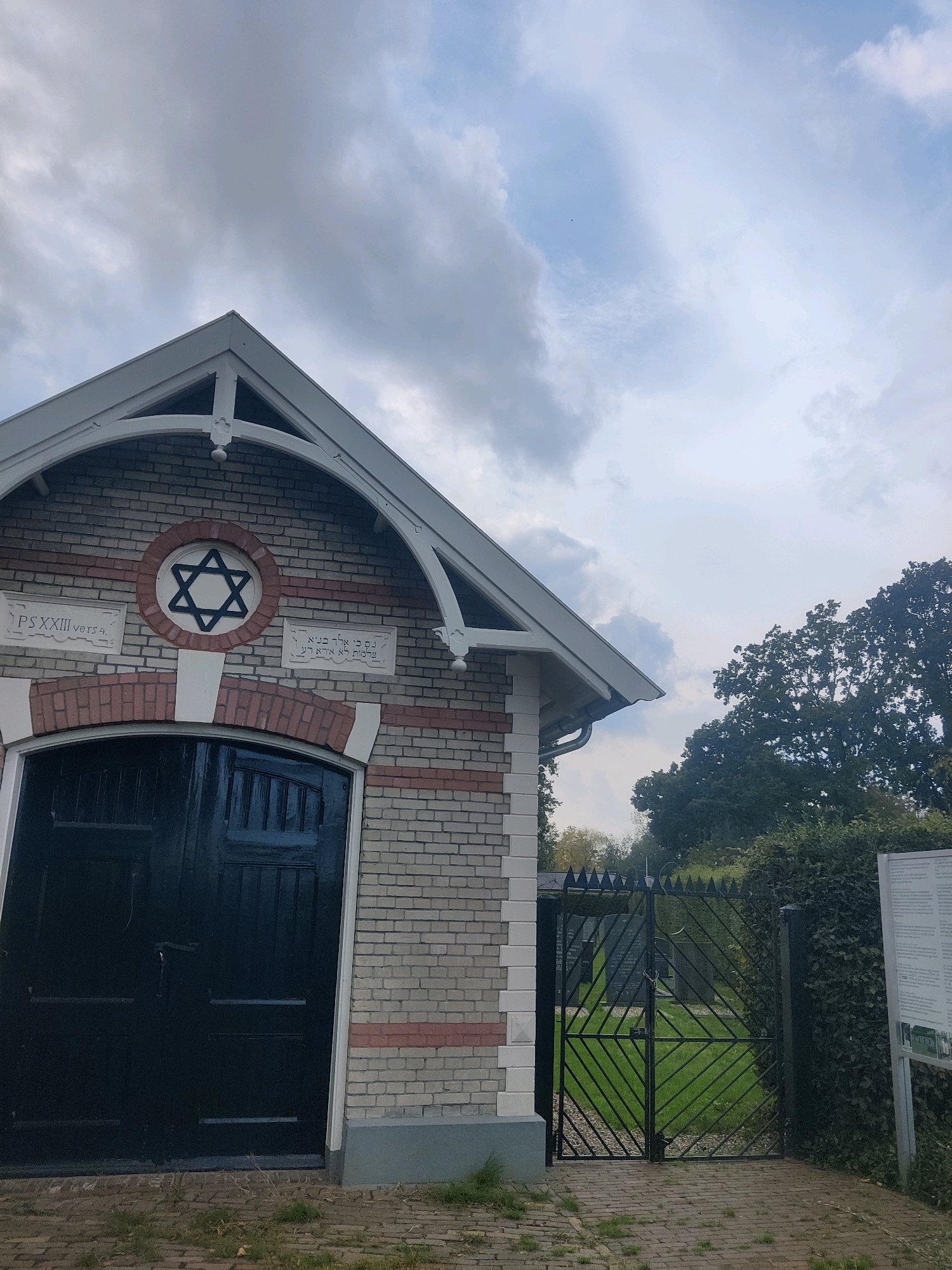 The image depicts the entrance to a Jewish cemetery in Winterswijk, Netherlands. The main focus is on a small, brick building with a prominent Star of David above a dark wooden door. The building has white trim and a gabled roof. To the right of the building is a black metal gate, also with pointed decorative elements, leading into the cemetery. Through the gate, tombstones are visible amongst green grass and trees. A sign with text and an image is posted to the right of the gate. The sky is overcast with clouds. The ground in front of the building and gate is paved with bricks.