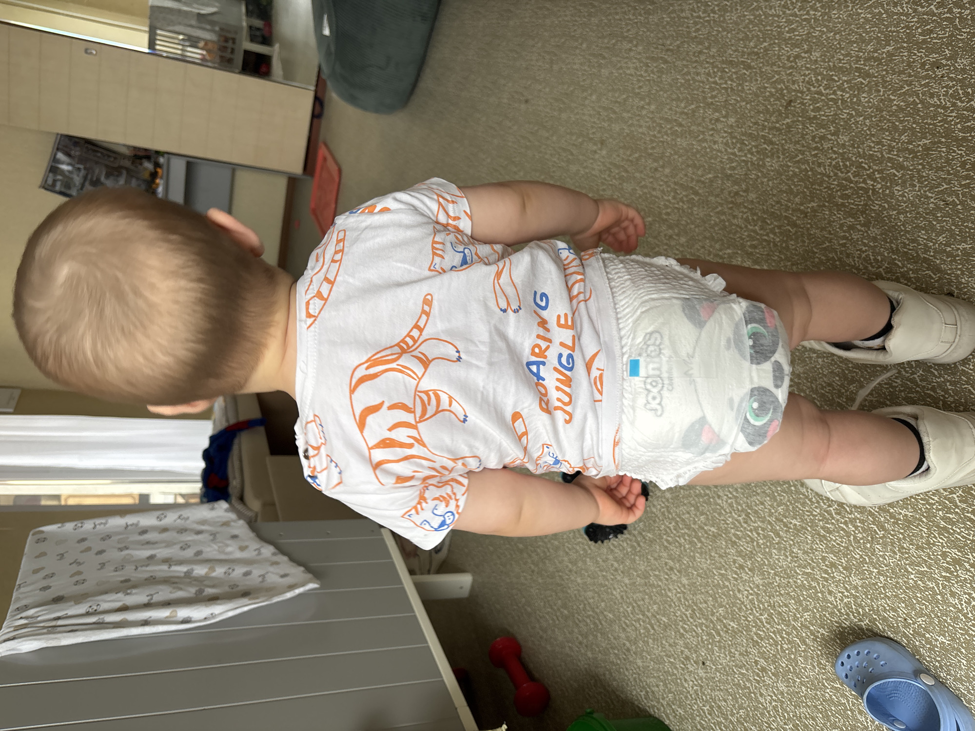 A young child, viewed from the back, stands facing away from the camera. The child is wearing a white t-shirt with orange tiger stripes and text that reads "ROARING JUNGLE," and a diaper featuring a panda design. The child is also wearing white sneakers. The environment appears to be an indoor residential setting. To the left, a grey crib is partially visible, with a white and patterned blanket draped over it. A red dumbbell and a green bucket are on the floor near the crib. To the right, a large dark green beanbag chair is visible. The floor is carpeted in a neutral tone with a textured pattern. In the background, a doorway with a white curtain and a mirrored wardrobe is present. The overall lighting suggests an indoor setting during daylight hours. The image has been captured in a residential apartment in Naberezhnye Chelny, Russia.