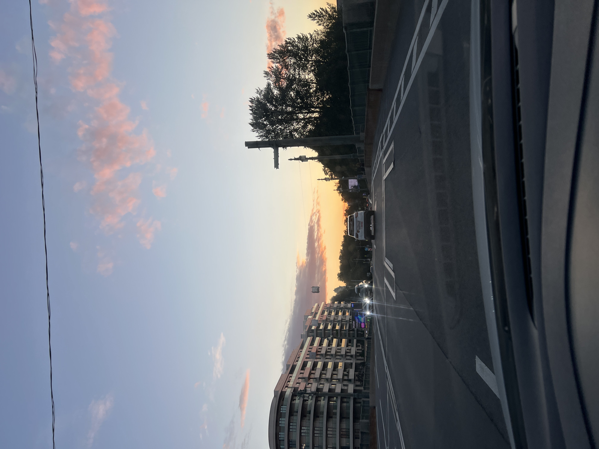 The image depicts a view from a vehicle driving on a road in Saint Petersburg, Russia, at dusk. The sky is a gradient of pale blue to soft orange, with scattered pink clouds. A power line stretches horizontally across the upper portion of the frame. To the left, a modern, curved residential building with multiple balconies is visible. On the right, silhouetted trees and a traffic signal structure are prominent. Several cars are on the road, with headlights illuminated. The road itself has white lane markings. The overall scene conveys a sense of an urban environment during the transition from day to night.