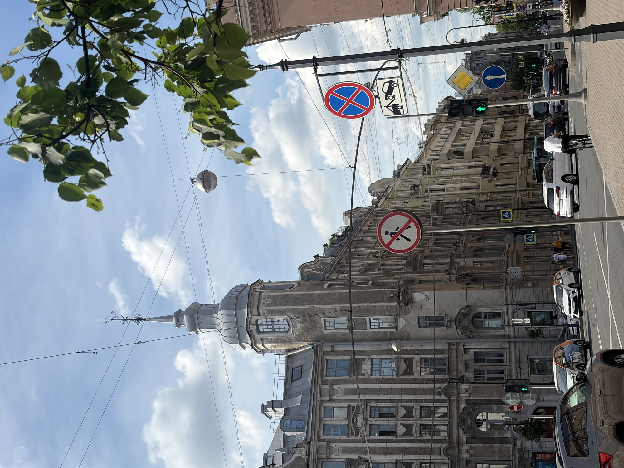 This image depicts a bustling urban street scene in Saint Petersburg, Russia, specifically at the intersection of Ulitsa Mayakovskogo and Nevsky Prospekt, prominently featuring the A.I. Khomyakov apartment building (often referred to as the "House with a tower") at Ulitsa Mayakovskogo 1. The scene is illuminated by daylight under a partially cloudy sky.
On the left, a multi-story historic building dominates, characterized by its grey, ornate facade with vertical pilasters, stucco detailing, and varied window designs. A distinctive corner tower, topped with a grey conical roof and a slender spire culminating in a finial, extends skyward. The building's mansard roof features several dormer and skylight windows. To the right, a continuous row of multi-story buildings, consistent with traditional European urban architecture, extends down the street; one building on the far right displays a reddish-pink facade.
The sky is bright blue with scattered white cumulus clouds. A network of overhead wires, likely for trams or electrical distribution, traverses the upper portion of the frame. A spherical street light fixture hangs from these wires in the upper center. A leafy green tree branch partially obscures the top right corner.
In the foreground and mid-ground, a multi-lane asphalt road is populated with various vehicles. The hood and front fender of a dark grey SUV are visible in the lower left. On the right side of the road, a person in a white t-shirt is riding a bicycle away from the camera. Other vehicles on the road include a white Audi Q5 SUV turning left, a white Kia Sportage, an orange-and-white Nissan Kicks (or similar compact SUV), and other cars and a white bus visible further down the street. Several pedestrians are discernible on the sidewalks and at a distant crosswalk.
Multiple street signs are present: a red circular 'no pedestrians' sign on the left; a red circular 'no stopping' sign with a 'tow-away zone' pictogram beneath it on the right; a yellow diamond 'priority road' sign; and a blue circular 'straight ahead only' sign. Traffic lights are visible, with green signals illuminated for forward movement. Black lamp posts and traffic signal poles are spaced along the street. A small green shrub is planted near the base of one building, and flower pots are visible on the right sidewalk further down. A clock face is mounted on the corner of a building on the right side.