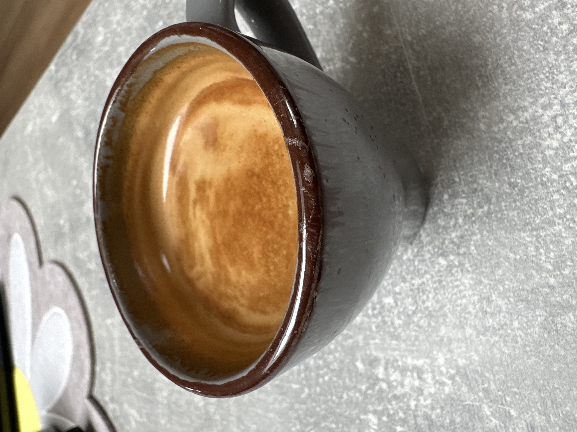 The image shows a close-up, eye-level view of a dark gray mug filled with a golden-brown espresso. The espresso has a crema layer with swirling patterns. The mug has a dark brown rim. The mug sits on a textured, gray surface. In the background, out of focus, are some light-colored objects on a yellow surface. The scene appears to be indoors, possibly in a cafe or home environment, but no specific location in Garmisch-Partenkirchen, Germany can be identified from the image. There are no people or discernible actions taking place.