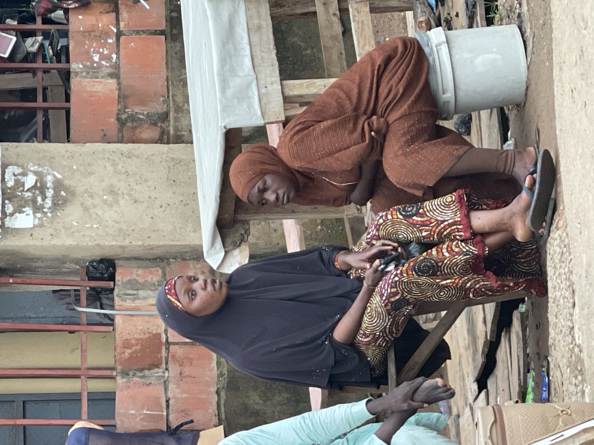 Two women are sitting on a wooden bench outdoors in Abuja, Nigeria. The woman on the left wears a black hijab and a patterned skirt. She is holding a mobile phone in her right hand. The woman on the right wears a brown hijab and a brown dress. She is holding her arms crossed in front of her. They are sitting next to a rough wooden structure covered with a white sheet. In the background, a wall made of reddish-orange bricks is visible. The ground is dirt and gravel, with some debris scattered around. A large white bucket is placed next to the woman on the right.