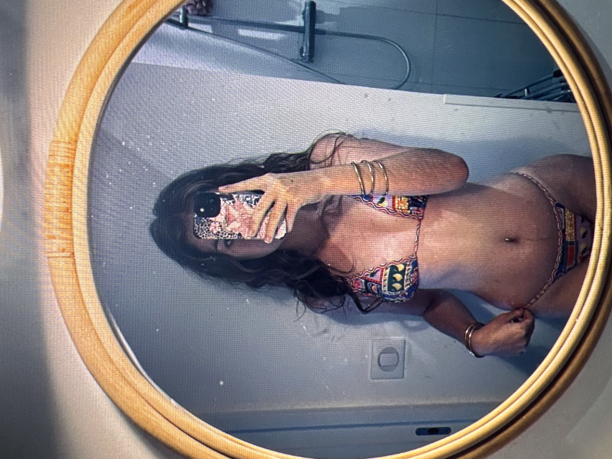 A person is taking a mirror selfie in what appears to be a bathroom. The person is wearing a colorful bikini and has long, dark, wavy hair. They are holding a phone with a floral case to take the picture. The mirror is framed in a round, light-colored wood. In the reflection, a white tiled wall is visible, along with a light switch and a shower head. The environment is indoors, likely a residential setting. There is no visual information in the image to determine the location in the city of Castelo Branco, Portugal.