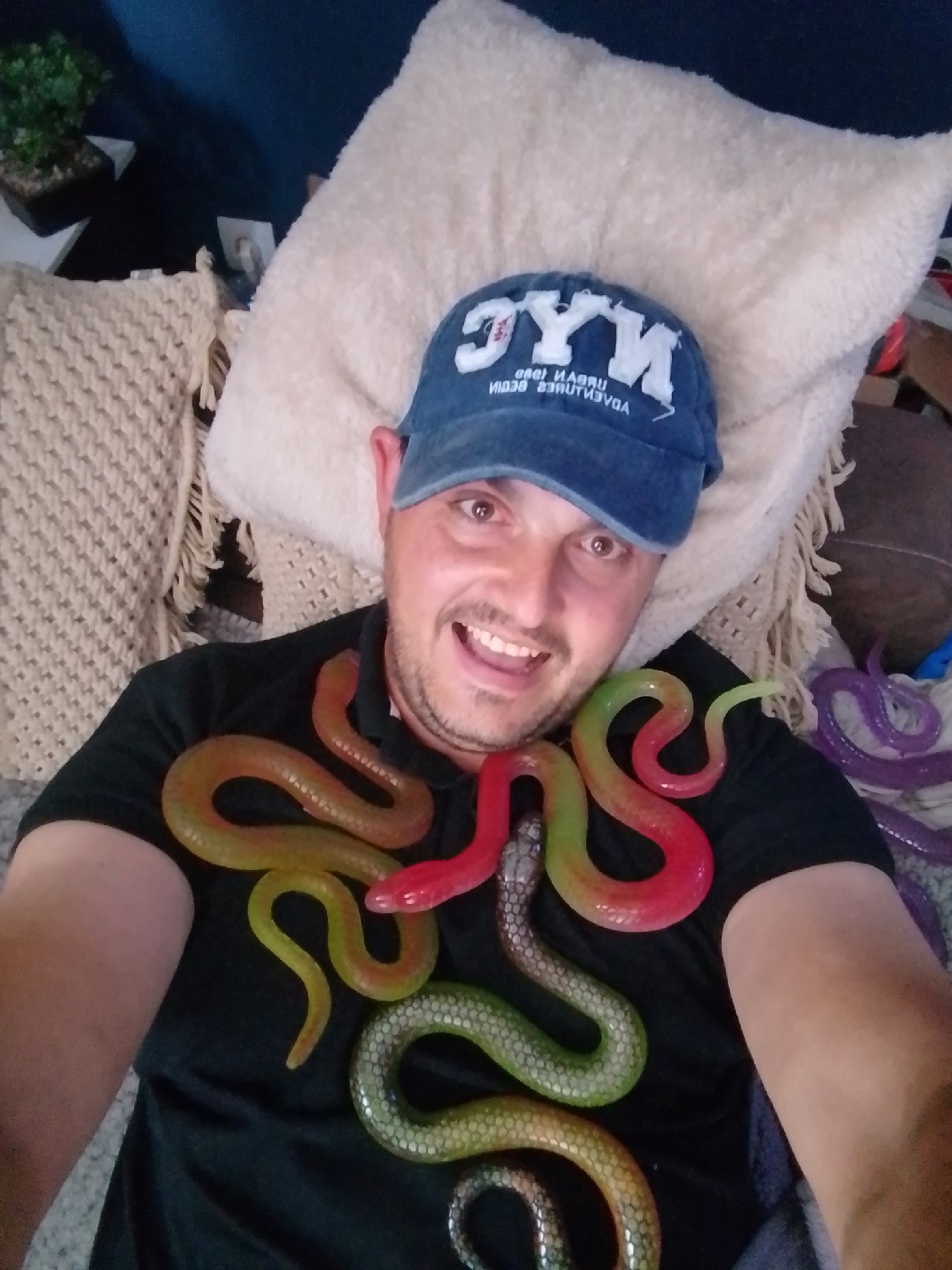 The image shows a man lying down, with his upper body visible. He is wearing a blue baseball cap with the letters "NYC" on it and a black t-shirt. Several brightly colored, segmented rubber snakes are draped around his neck and chest, in shades of red, orange, yellow, green, and purple. The man is smiling. He appears to be indoors, possibly on a couch or bed, with pillows and textured fabric visible in the background. There is a small potted plant in the upper left corner. The scene is not located in Wedel, Germany.