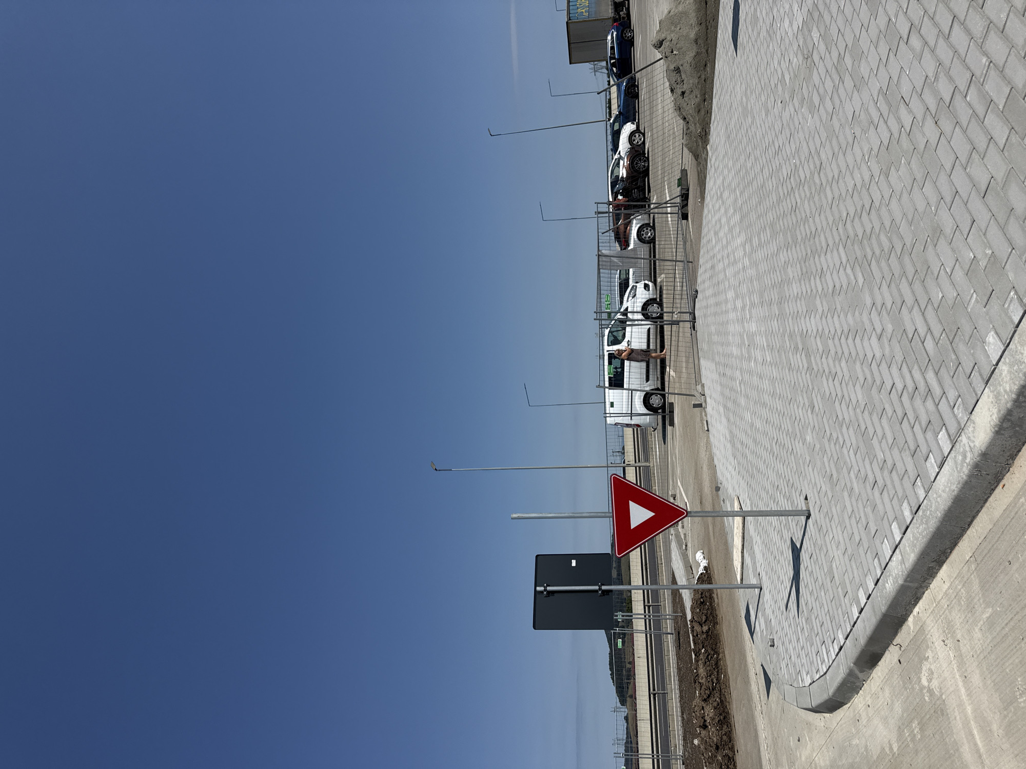 In Sanpetru, Romania, a clear, bright day unfolds with a vast expanse of deep blue sky dominating the upper portion of the image. The lower half of the frame presents a scene of ongoing development or a recently completed public space. In the foreground, a paved area made of interlocking grey bricks forms a curved section, meeting a concrete curb. To the left, a patch of rough, disturbed earth indicates construction or landscaping activity. A prominent red and white yield sign, a triangular traffic marker, stands on a metal post. Beside it, a plain black rectangular sign on a similar post is also visible. Beyond this immediate area, a grey metal fence separates the scene from a road or thoroughfare. Several vehicles, including a white van and a dark blue car, are parked behind the fence, some partially obscured. A person wearing a light-colored top and dark trousers stands near the white van, facing away from the camera. Further in the background, more cars and a large white truck with a trailer are visible, also behind fencing. Distant rolling hills and power lines are faintly discernible on the horizon under the expansive sky.