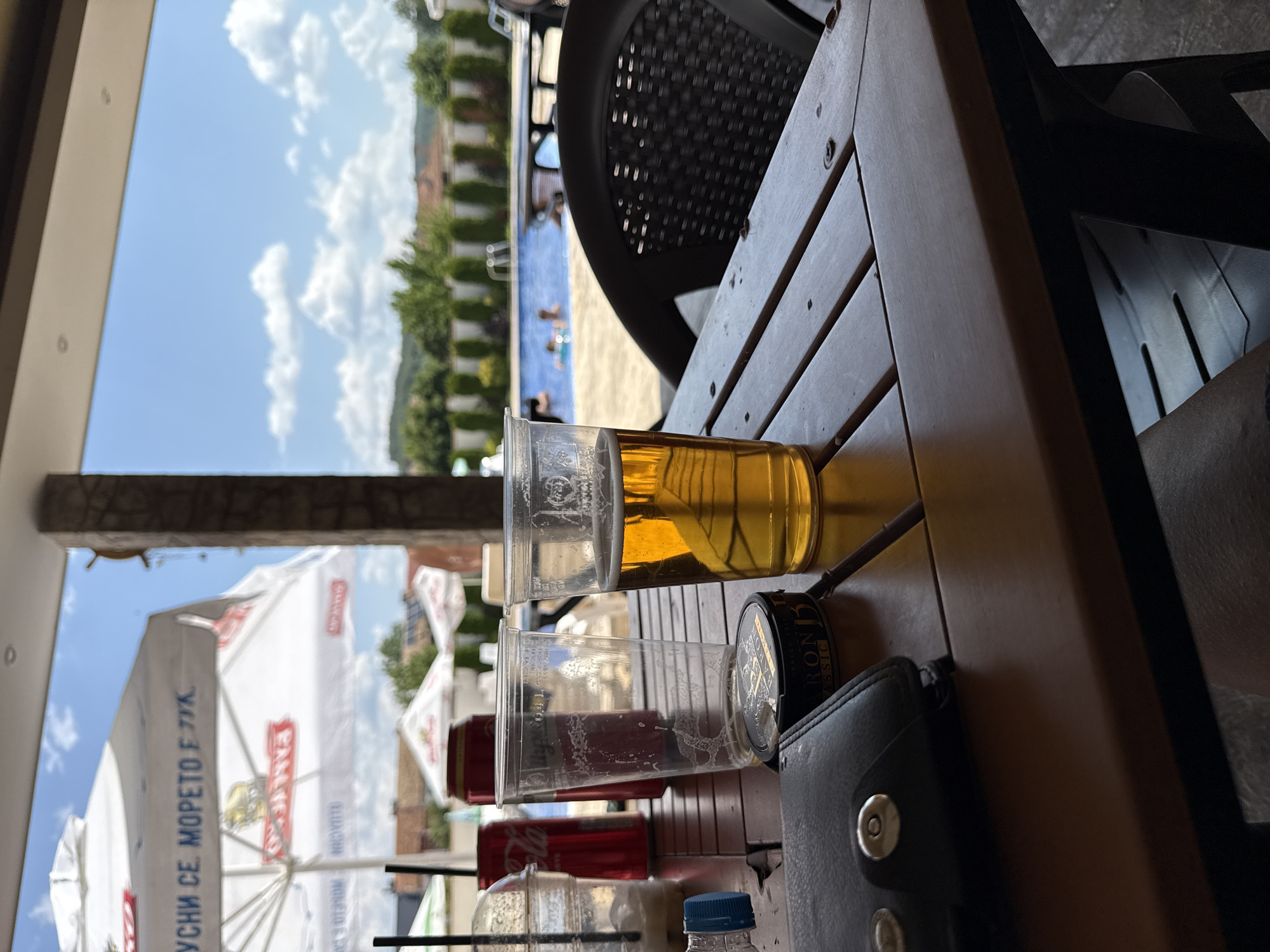 The image depicts a close-up view of a wooden table outdoors, set against a backdrop of a swimming pool and surrounding greenery. In the foreground, there are several objects on the table. To the left, a black leather wallet with silver button closures is partially visible. Next to it is a round, dark tin container with gold lettering that appears to be tobacco. In the center, two clear plastic cups are present. One is empty with condensation marks, and the other contains a golden-colored liquid, likely beer, with a frothy head. A red aluminum can is also visible behind the empty cup. In the background, a swimming pool with a few people can be seen, along with lounge chairs and umbrellas. The environment suggests a relaxed, possibly resort-like setting. The context provided indicates this scene is located in the city of Strazhitsa, Bulgaria.