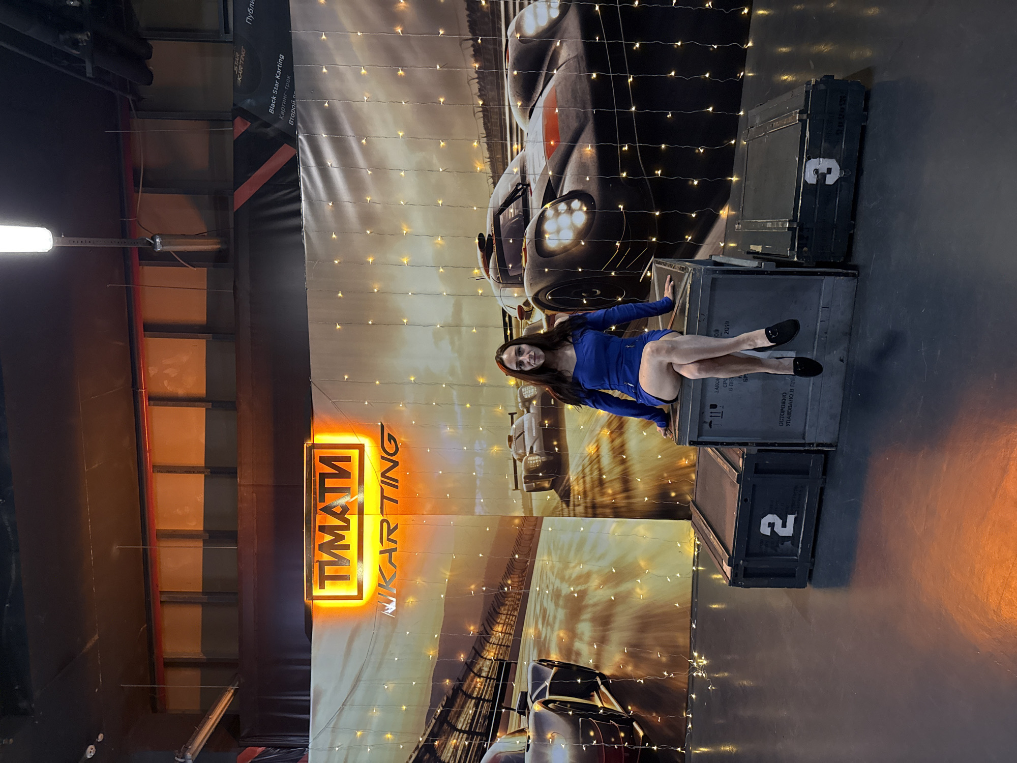 A woman in a blue dress is sitting on a crate in front of a backdrop of racing cars and a "TIMATI KARTING" sign. The backdrop is adorned with string lights. There are three crates arranged in a podium formation, with the woman sitting on the middle crate. The crate on the left has the number "2" on it, and the crate on the right has the number "3" on it. The floor is dark and reflective, indicating a polished surface, possibly a studio or event space. The overall impression is a staged photo opportunity related to karting.
