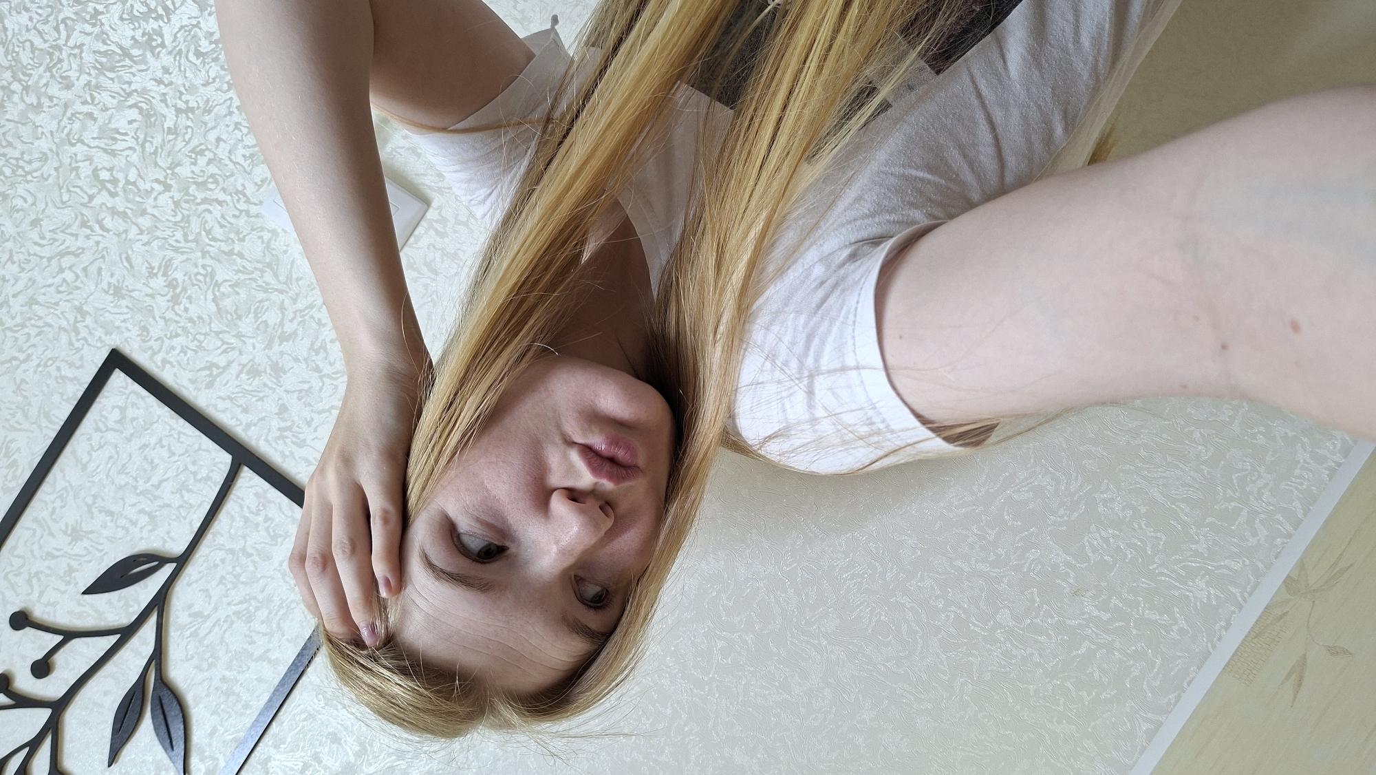 A young woman with blonde hair is taking a selfie. She is wearing a white t-shirt. Her left arm is extended, holding the camera. Her right hand is on her head, near her temple. She is looking up and to the right, with her lips pursed. The background is a textured, cream-colored wall with a decorative black frame containing a branch motif. The scene appears to be indoors, possibly in a residential setting. There is no indication of the location being in the city of Большой Букор, Russia.