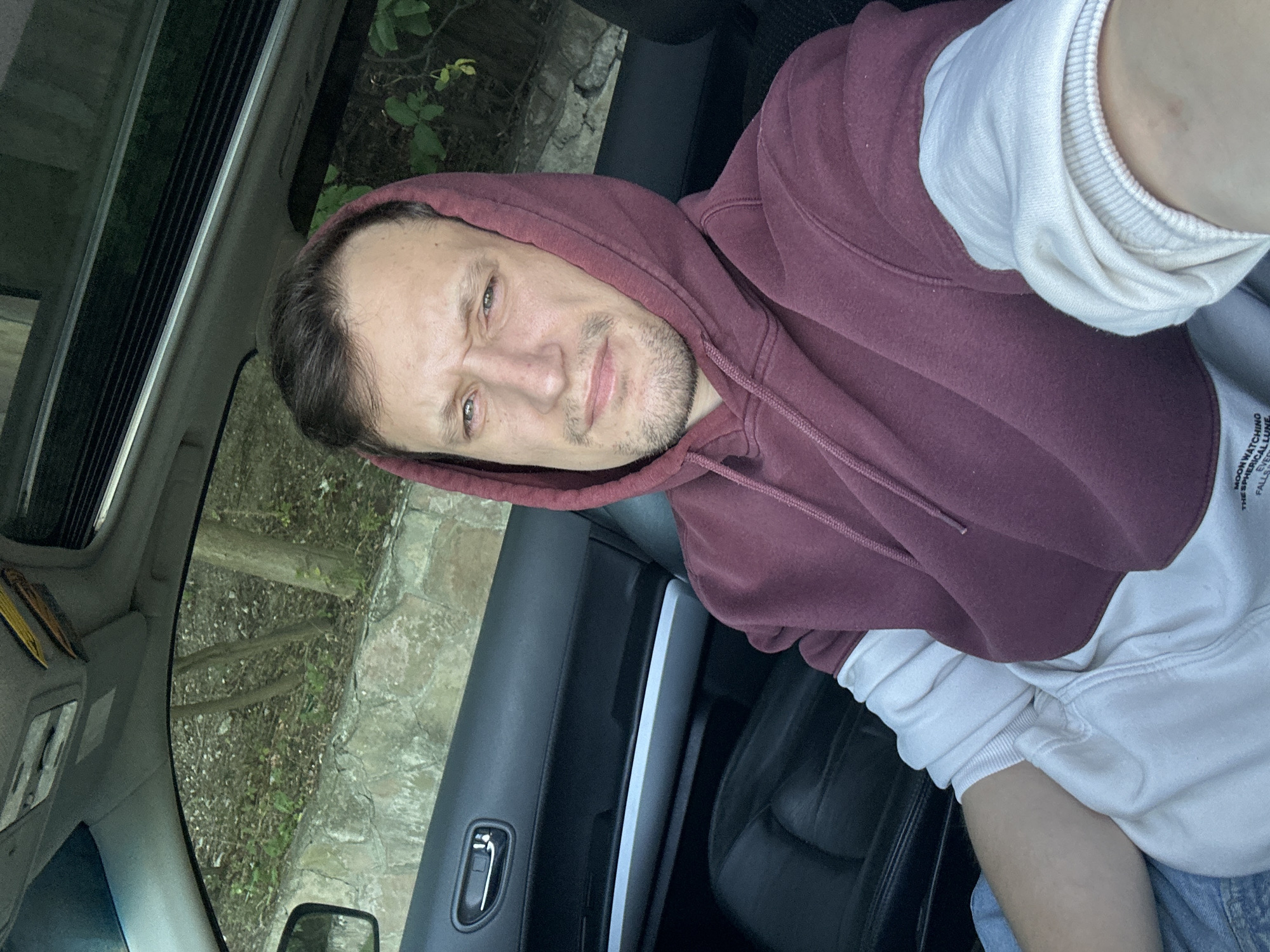 A man with short brown hair and stubble is sitting in the driver's seat of a car. He is wearing a maroon hooded sweatshirt over a white long-sleeved shirt and blue jeans. The car's interior is visible, including the black leather seats, door panel, and the sunroof. Outside the car, a stone wall and trees are visible. The man is looking directly at the camera with a neutral expression. The location is implied to be Makiv, Ukraine, although this is not explicitly visible in the image.