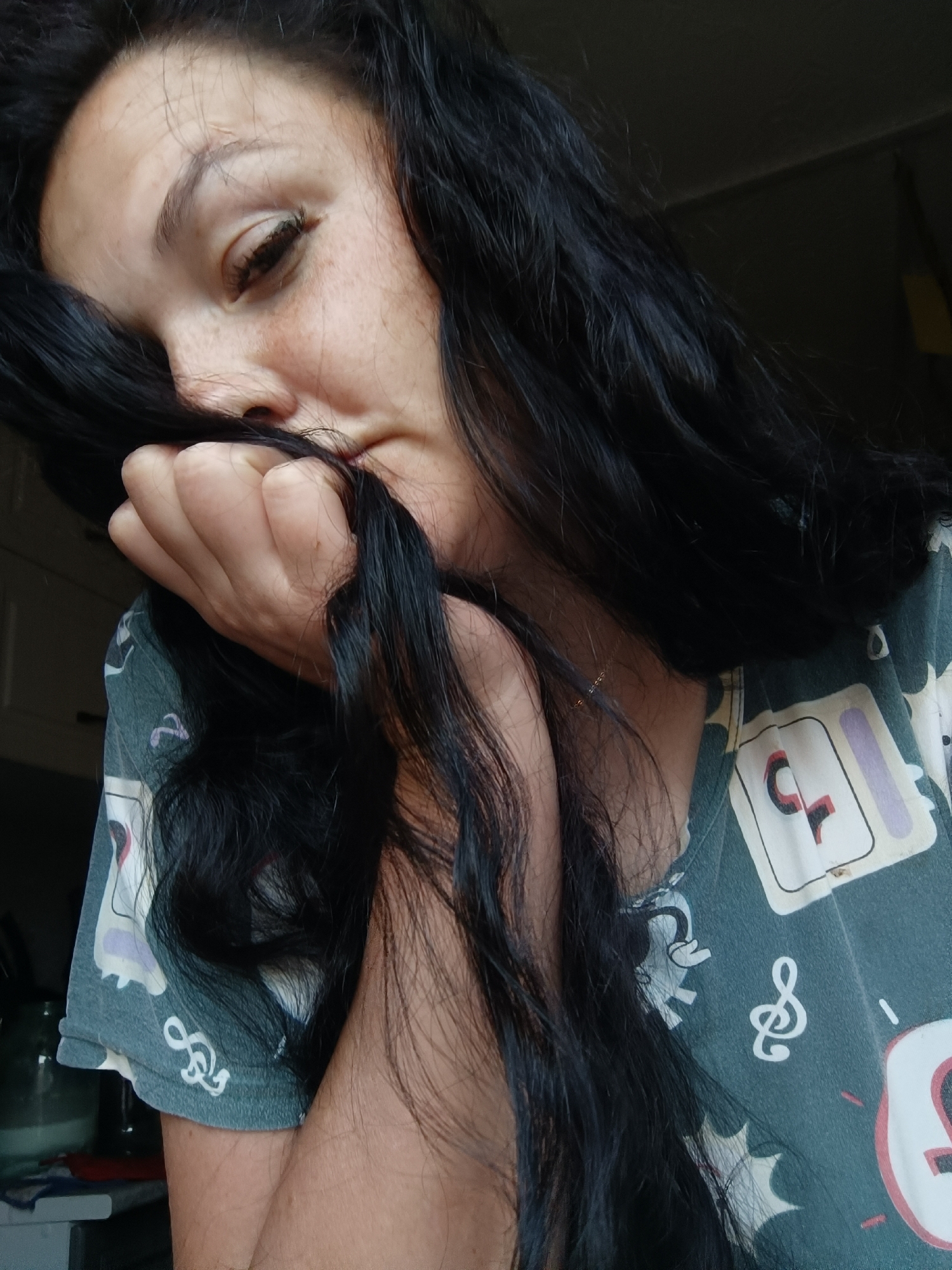 A woman with long, dark hair is partially obscured by her hair, which she is holding with her right hand. Her eyes are closed, and she appears to be leaning into her hair. She is wearing a dark green t-shirt with a repeating pattern of musical notes and cartoonish figures. The background is dark and out of focus, with hints of cabinets and a jar. The lighting suggests an indoor setting, possibly a home. The image is a close-up shot, focusing on the woman's face and upper body. There is no discernible information in the image that would indicate the location in the city of Karsha, Russia.