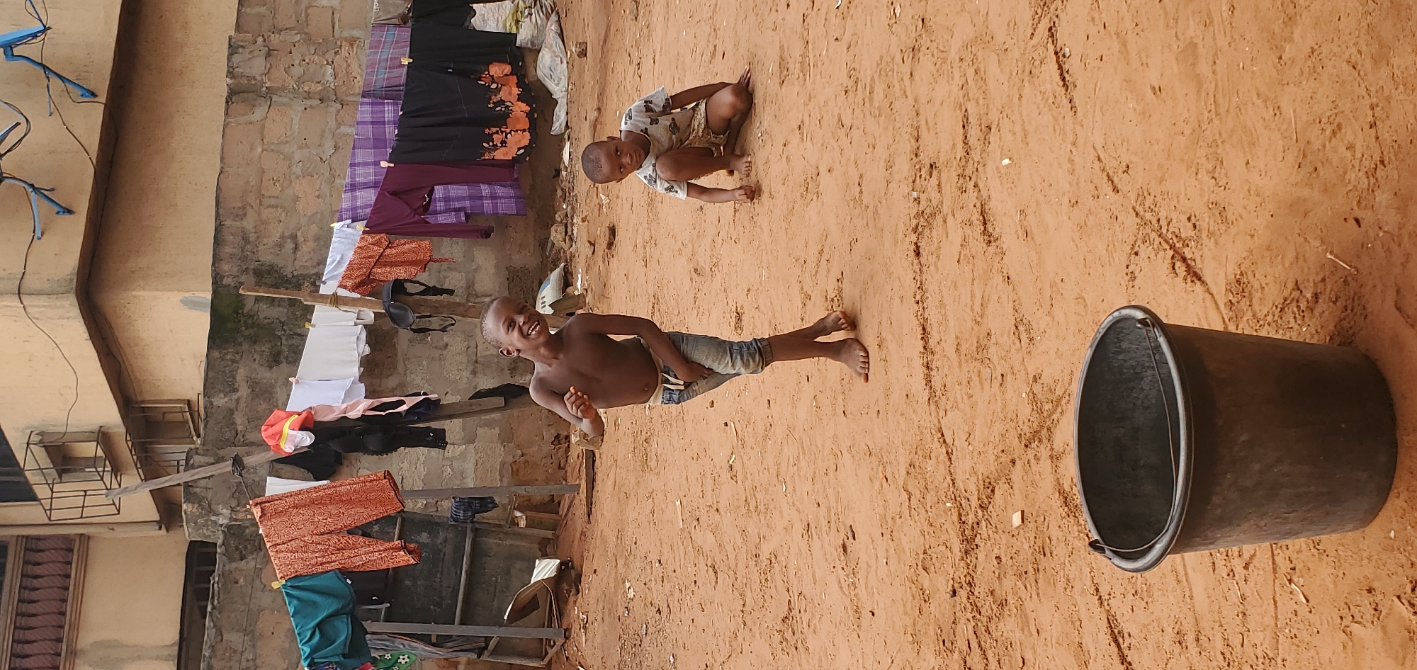 The scene captures two children playing in a sandy yard in Onitsha, Nigeria. One child is standing, shirtless and wearing jeans, with a wide smile. The second child sits in the sand, observing. Laundry is hanging on a line attached to a weathered building. A dark bucket sits nearby on the sandy ground.