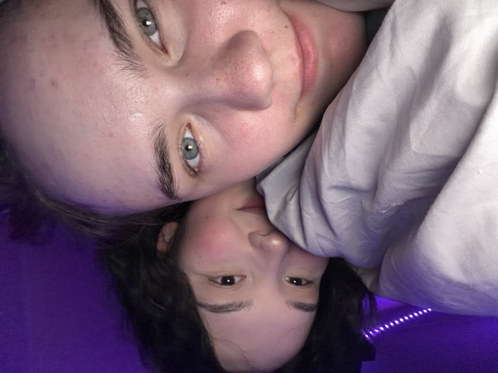 A close-up, eye-level photograph features two individuals, likely young adults, in a dimly lit indoor setting. The foreground individual, positioned at a slight angle with their head tilted right, looks directly into the camera. This person exhibits fair skin with scattered freckles, distinctive light blue-green eyes, and parted pink lips forming a subtle smile. Dark hair frames their face.
Behind and slightly to the left, the second individual's face is partially visible, primarily their eyes and upper facial features, as they rest their head near the first. This individual has dark brown eyes and dark, curly hair. A soft, light-colored fabric, likely a blanket or duvet, covers both individuals, with a portion of it obscuring the lower half of the background person's face.
The background consists of a plain, dark purple wall, brightly illuminated by a visible string of purple LED lights in the upper left quadrant. The overall lighting scheme is dominated by a purple hue.