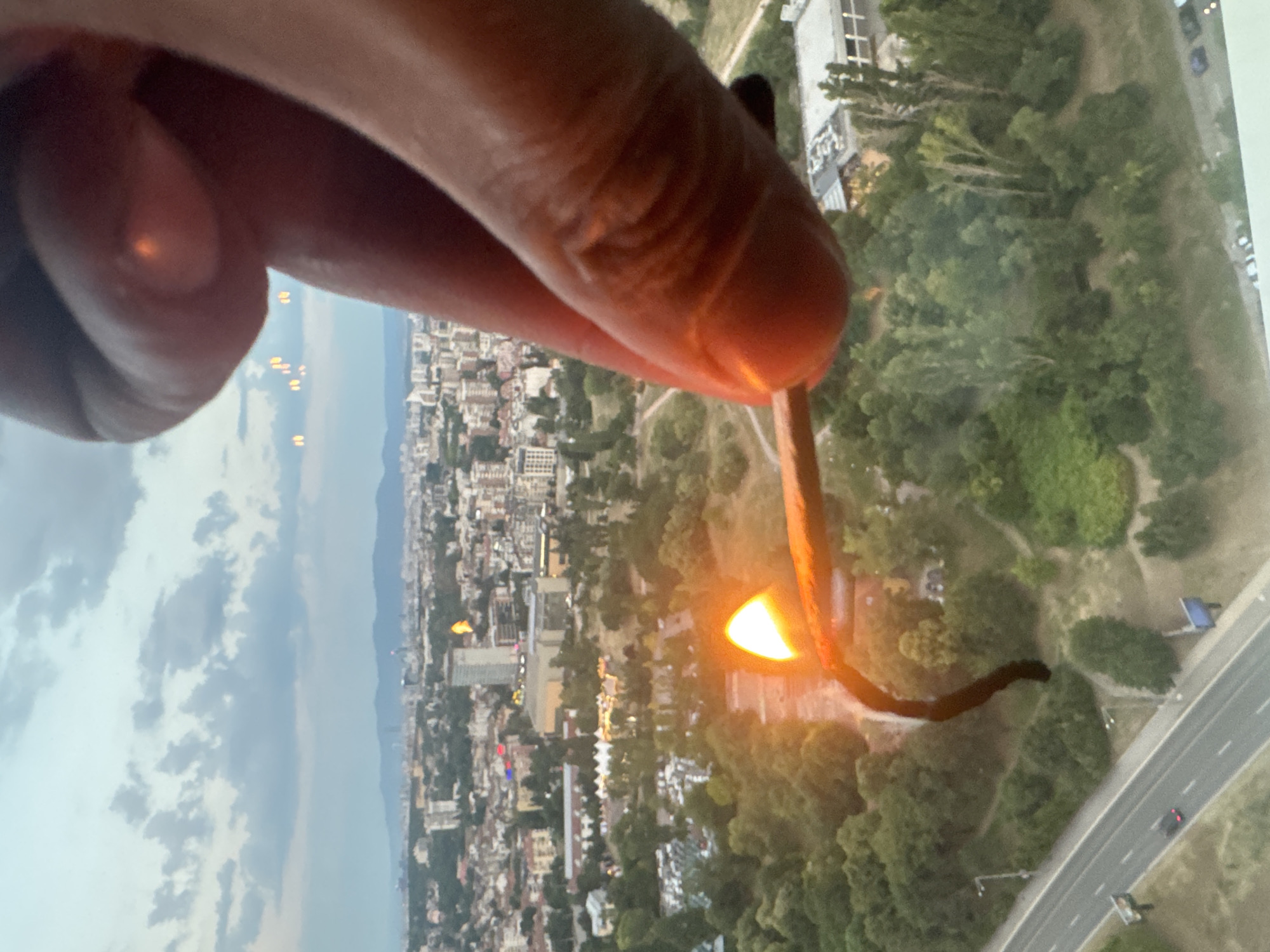 In Sofia, Bulgaria, a close-up shot focuses on a hand holding a lit match. The matchstick is mostly obscured by the flame, which emits a bright orange glow. The background reveals a panoramic view of the city, characterized by a dense collection of buildings, green spaces, and distant hills under a cloudy sky. A road with a car is visible in the foreground, suggesting an urban setting. The lighting conditions indicate it is likely dusk or dawn.