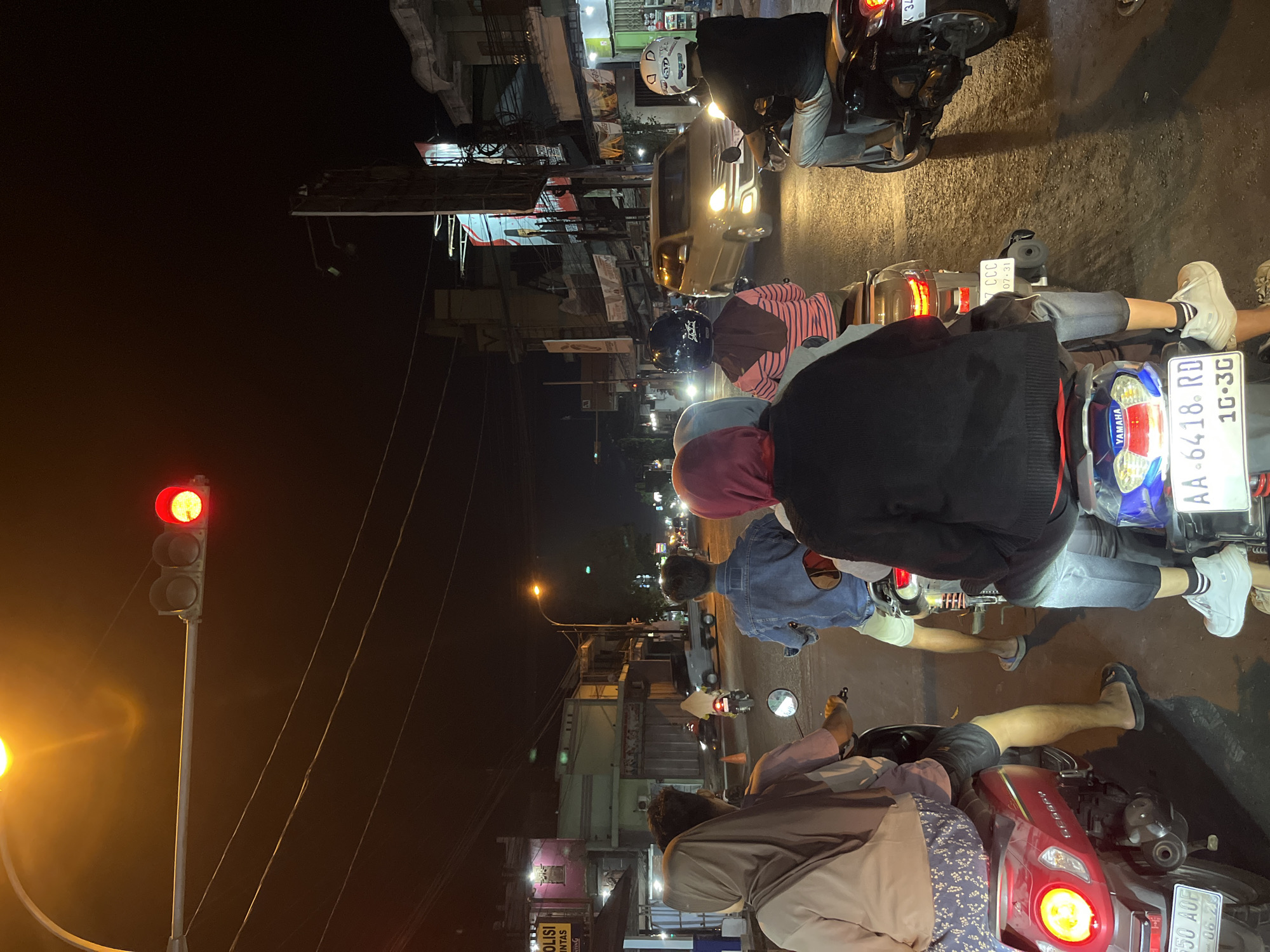 In Kalinyamatan, Indonesia, at night, a group of people are on motorbikes at a traffic light. The traffic light is red. The scene is a street with buildings on either side. There are other vehicles on the road, including cars and other motorbikes. The people are wearing casual clothing and some are wearing hijabs. The atmosphere is busy and urban.