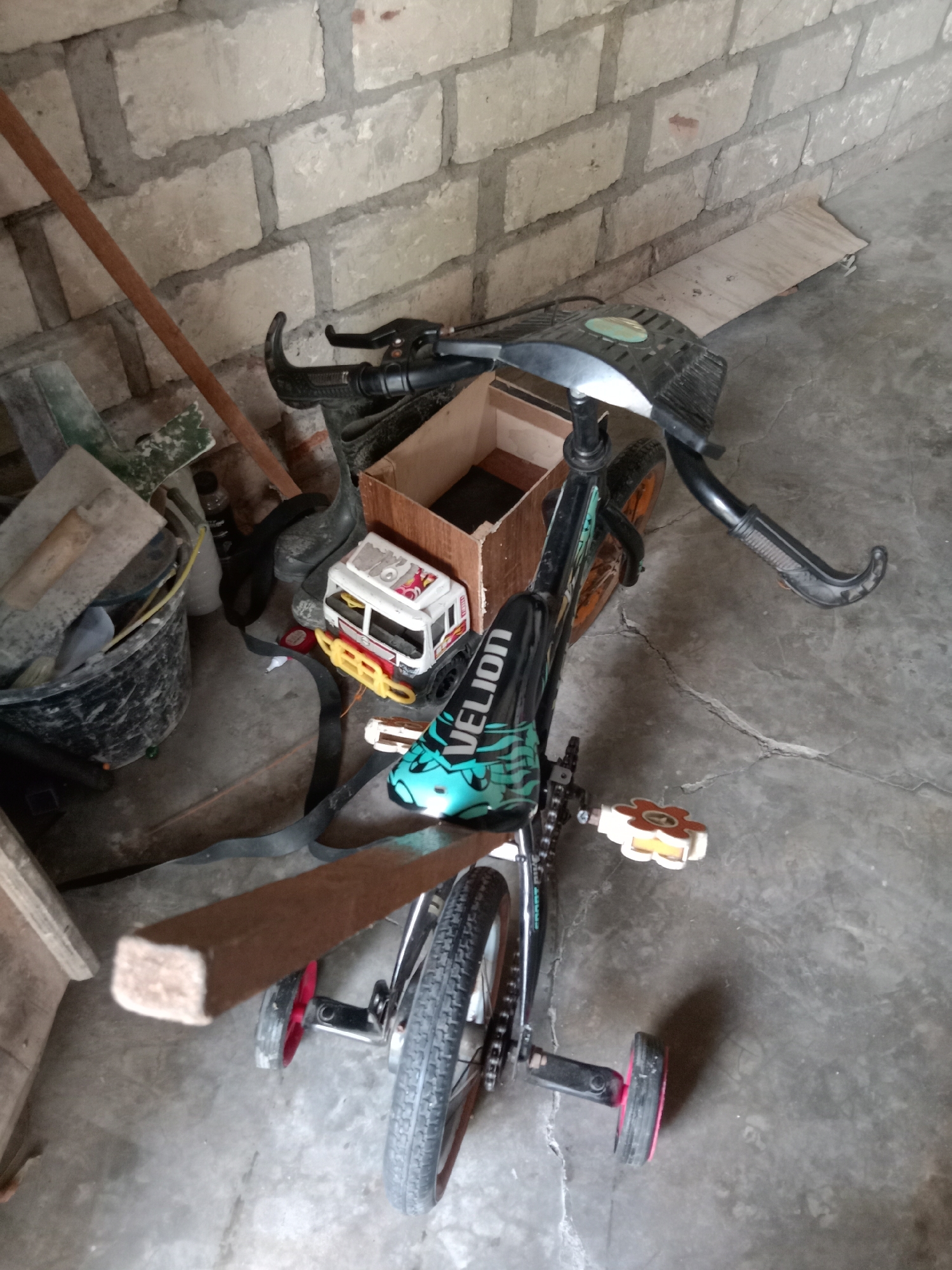 The image depicts a tricycle, likely a child's, propped against a brick wall in what appears to be an outdoor storage area or shed. The tricycle is black with teal accents on the seat and handlebars. It has training wheels. Scattered around the tricycle are various items, including a toy truck, a cardboard box, a black bucket filled with what appear to be tools or cleaning supplies, and a wooden plank. The ground is concrete and shows some cracks. The brick wall is constructed from light-colored bricks with visible mortar lines. The overall impression is one of a cluttered, utilitarian space. The specific location within Jumput, Indonesia, is not discernible from the visual cues in the image.