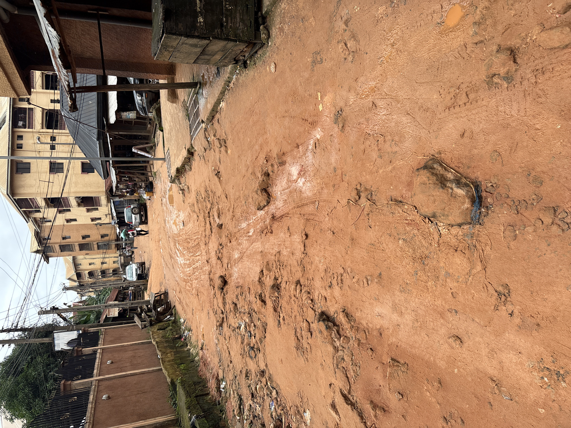 The image depicts a street in Nkpor, Nigeria, characterized by a significant amount of red mud and debris. The road surface is uneven, with tire tracks and footprints visible in the mud, indicating recent vehicular and pedestrian activity. Several cars are parked or driving on the road, some appearing to be utility vehicles. Buildings line both sides of the street, with some exhibiting signs of wear and tear. The architecture suggests a mix of residential and commercial spaces, with some shops featuring open fronts and makeshift structures. Electrical wires crisscross the sky, typical of urban environments. A large wooden crate is visible on the right side of the frame. The overall scene conveys a sense of urban infrastructure that is in a state of disrepair, likely exacerbated by recent rainfall.